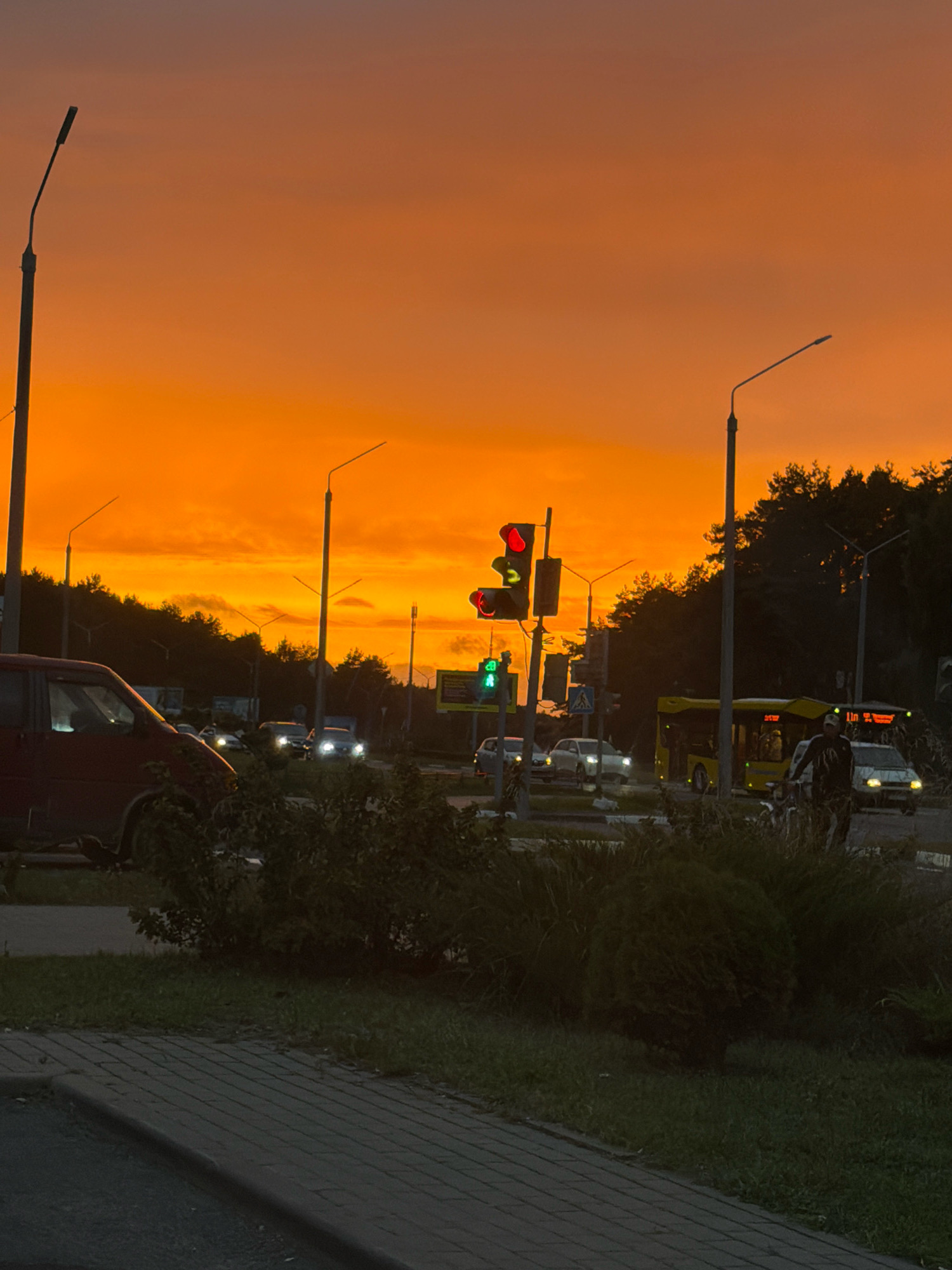 The image captures a sunset scene in Salihorsk, Belarus, characterized by a vibrant orange sky. In the foreground, a dark-colored van is parked on the left, with its wheel and part of the passenger side visible. Adjacent to it, dense green foliage and grass cover the ground, bordering a paved walkway made of gray bricks.
The middle ground features a traffic intersection. A traffic light displays a red signal, with the green signal also illuminated. Several cars and a yellow bus are visible on the road. A person on a bicycle is also present, moving towards the right side of the frame. Streetlights stand tall along the road, some with visible lamps. Trees and dense bushes line the background, silhouetted against the dramatic sky. The overall atmosphere suggests an urban setting at dusk.