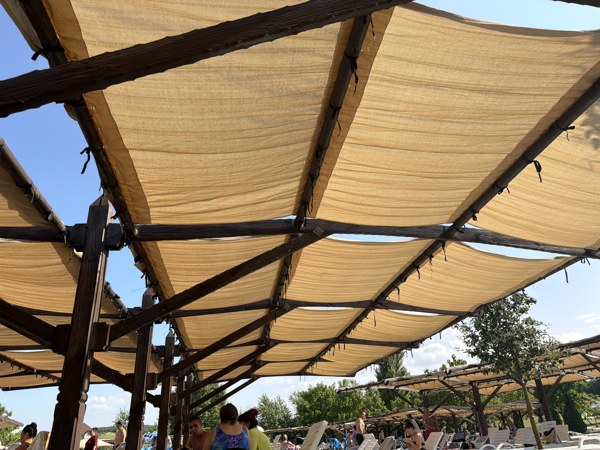 The image depicts an outdoor area with a large, wooden-framed canopy covered by tan fabric, providing shade. The canopy structure is elaborate, with multiple beams and supports. Beneath the canopy, several people are visible, some seated on lounge chairs, suggesting a recreational setting such as a resort or park. Trees and a clear blue sky are visible in the background, indicating an outdoor environment. The scene is likely located in Sociteni, Moldova, as this is a known location for such outdoor leisure facilities.