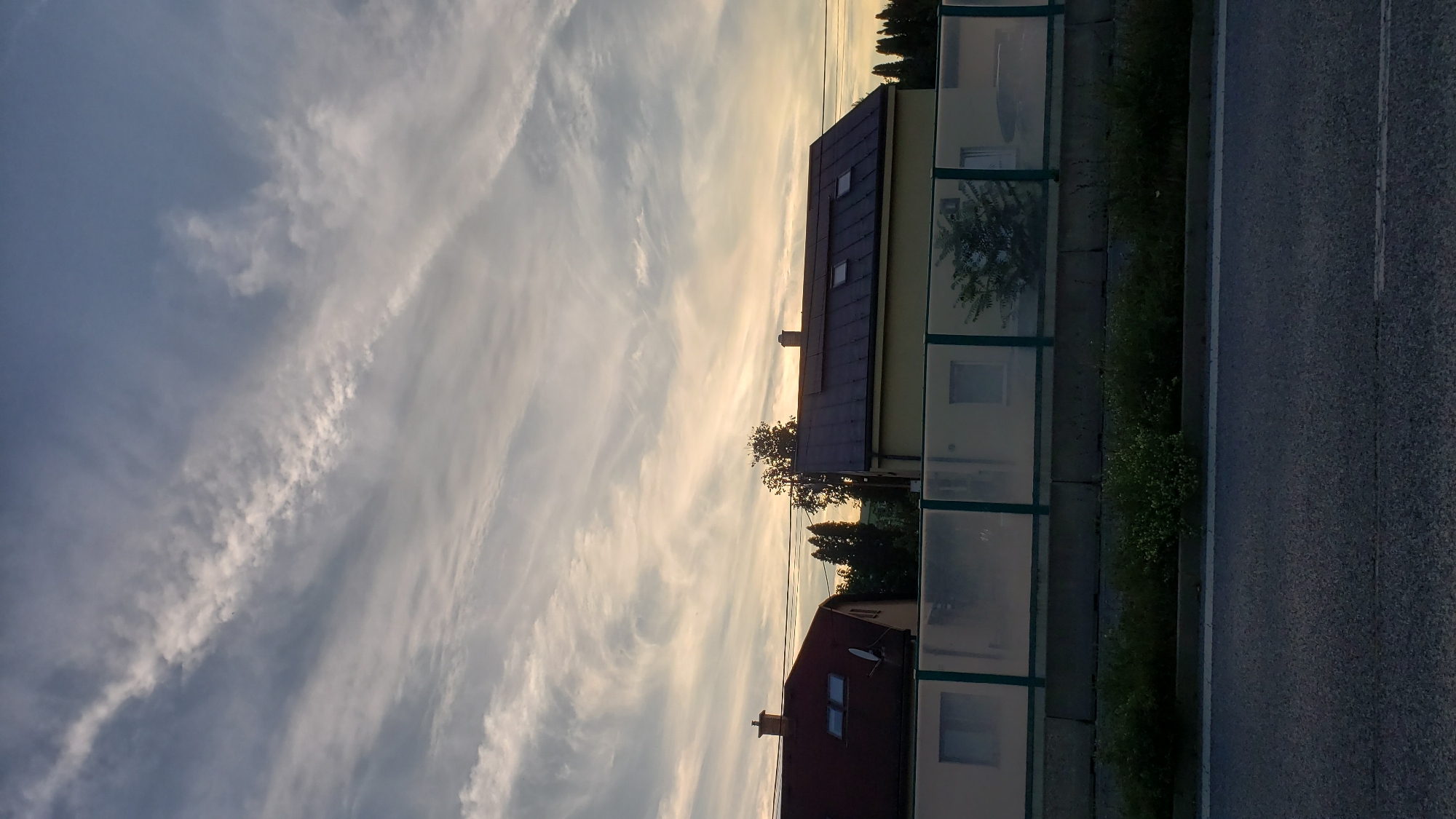 The image shows a roadside view in Prešov, Slovakia, with a cloudy sky at sunset. In the foreground, a dark asphalt road is visible. Behind the road, there is a strip of green vegetation and a concrete barrier. Beyond the barrier, there is a modern sound barrier made of translucent panels with green framing. Two houses are partially visible above the sound barrier. The house on the left has a dark red roof with two skylights and a chimney. The house on the right has a dark roof with two skylights and solar panels. Trees are visible behind the houses. The sky is filled with wispy, elongated clouds, illuminated by the setting sun, creating a dramatic effect. The overall scene suggests a residential area adjacent to a road, possibly a highway or a main thoroughfare.