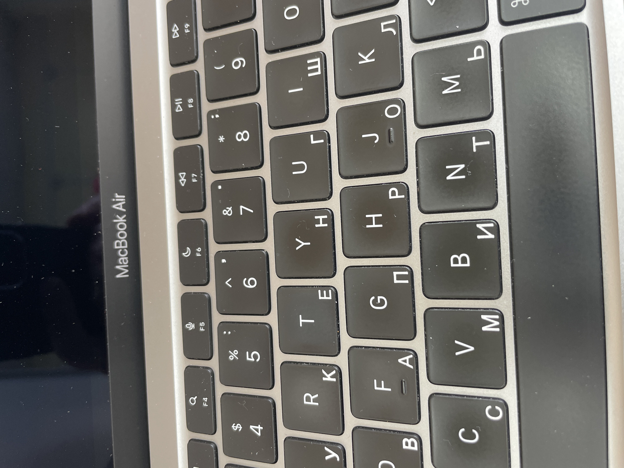 The image shows a close-up of a MacBook Air keyboard. The keyboard is black with white lettering. Some keys have Cyrillic characters in addition to Latin characters. The MacBook Air logo is visible above the keyboard. The image is taken from a slightly elevated angle, looking down at the keyboard. There is no indication of the location being Moscow, Russia. The environment appears to be indoors, likely an office or home setting, with a portion of the laptop screen visible at the top. No people or interactions are present in the image.