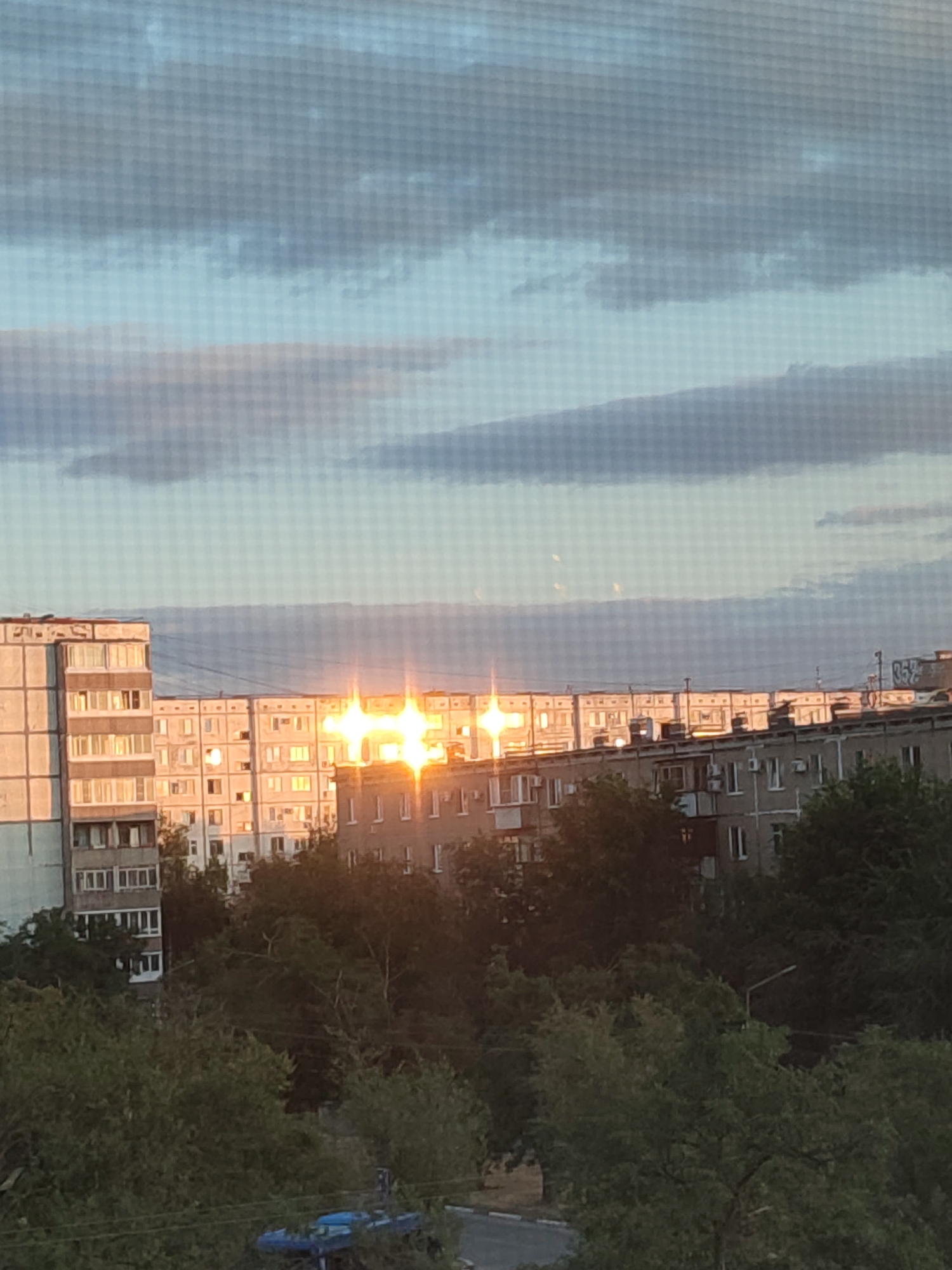 The image depicts a residential area in Volzhsky, Russia, with a bright sun reflecting off the windows of multi-story buildings. The architecture suggests Soviet-era construction, characterized by uniform apartment blocks. Dense foliage from trees obscures the lower sections of the buildings and extends into the foreground, indicating a green and possibly park-like environment surrounding the residential blocks. The sky is partly cloudy, with the sun creating a glare that emphasizes the reflective surfaces of the windows. The overall impression is a typical urban landscape during daylight hours, with a focus on the interplay of sunlight and architecture within a natural setting.