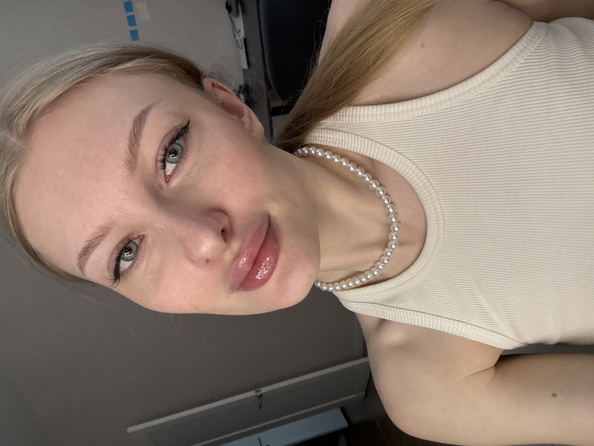 This is a close-up, medium shot, portrait of a young Caucasian woman with blonde hair pulled back into a ponytail. She is wearing a cream-colored ribbed tank top and a pearl necklace. The woman has light skin, green eyes, and full, glossy lips. She is looking directly at the camera. The background is dimly lit and appears to be an indoor setting, possibly a room in an apartment, with a dark wall and a doorway visible on the left. There is no indication of the city of Perm, Russia, or any other specific geographic location.