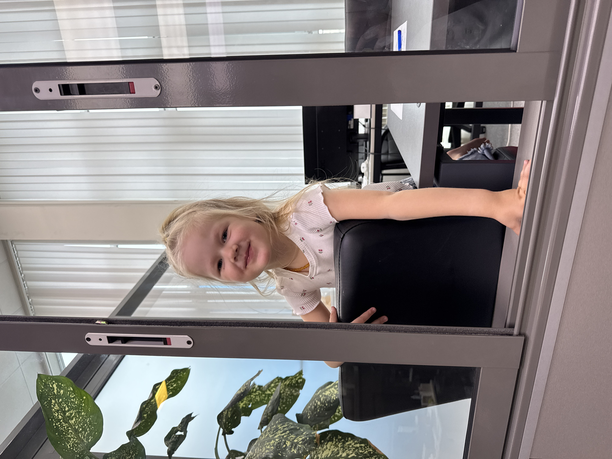 A bright, indoor portrait features a smiling young blonde-haired child peering over a grey-framed glass partition. The child, with fair skin, wears a light-colored ribbed t-shirt adorned with small red cherry patterns and a subtle beaded necklace, looking directly at the camera with a happy expression.
On the child's side of the partition, a dark chair or surface is visible, with their right arm resting upon it. The background behind the child, visible through the clear glass, shows an interior space resembling an office, featuring a desk, another chair, and white horizontal blinds on a distant window.
On the camera's side of the partition, a vibrant green and yellow variegated plant is seen through the lower glass pane, with a clear blue sky visible beyond it, indicating daytime. The grey partition frame has a silver-colored locking mechanism near the top, displaying a red indicator. The setting suggests a modern indoor environment, possibly an office or public building in Perm, Russia.