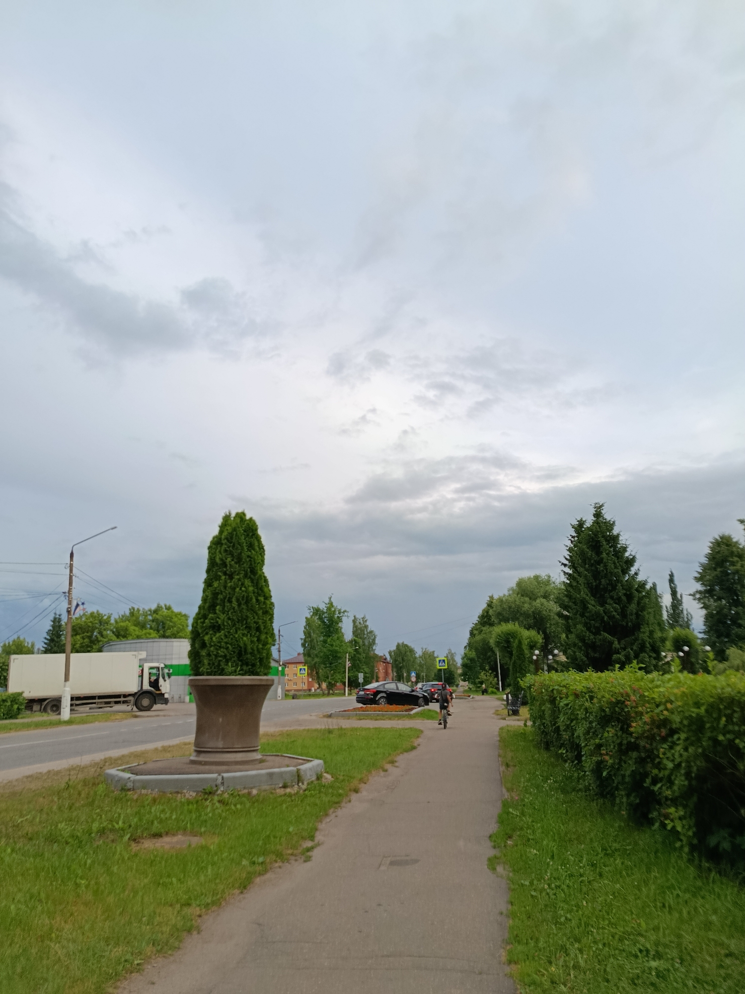 The image shows a street scene in the city of Vereya, Russia, under an overcast sky. In the foreground, a paved sidewalk runs along a grassy area. A large, urn-like structure stands on a concrete base in the grassy area. Further down the sidewalk, a person is riding a bicycle. On the street to the left, a white truck is parked. Across the street, there are trees and buildings, including a yellow building with red accents. The background features a line of trees and more buildings. The overall scene depicts a quiet, suburban or semi-urban environment.