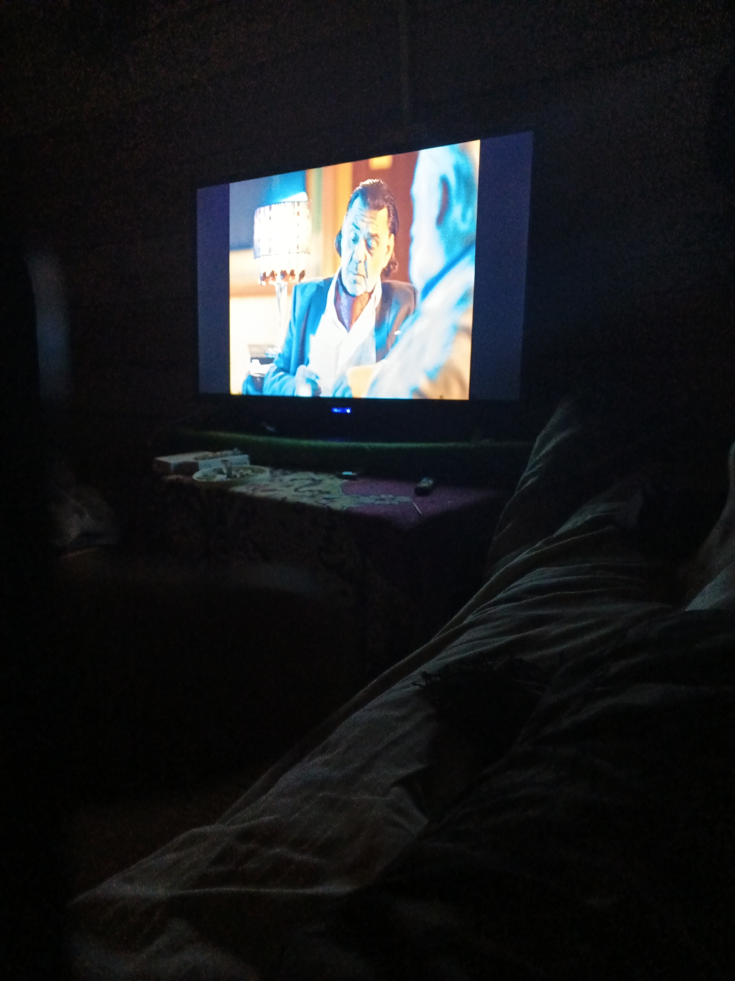 The image shows a television screen displaying a scene with two men. The man on the left is wearing a suit jacket over a white shirt, while the man on the right is wearing a light-colored shirt. They appear to be indoors, possibly in a room with dim lighting. In the foreground, there are blankets or bedding, suggesting someone is lying down while watching the television. The setting appears to be a domestic environment. There is no identifiable information in the image to determine the location in Strelka, Russia.