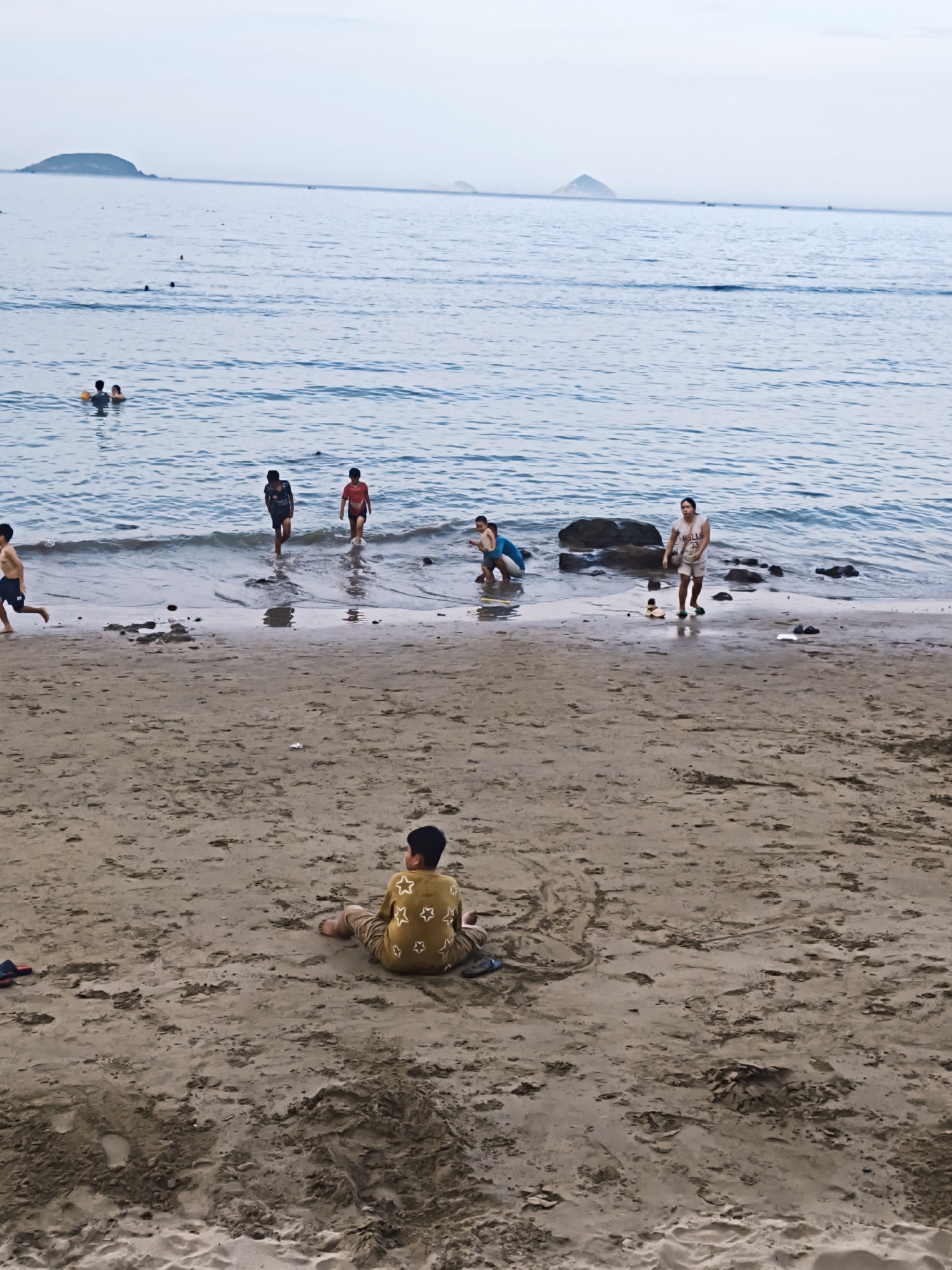 The image depicts a beach scene in Nha Trang, Vietnam, with the ocean stretching to the horizon under a pale sky. Several individuals are visible on the shore and in the shallow water. In the foreground, a young boy is sitting on the sand, facing away from the camera, wearing a yellow shirt with white stars. Further down the beach, near the water's edge, a woman in a white tank top and shorts is standing, and a child in a blue swimsuit is partially visible. Several other children are in the water, some wading and others playing. Distant islands can be seen on the horizon. The sand is wet and textured, with footprints and debris scattered across it.