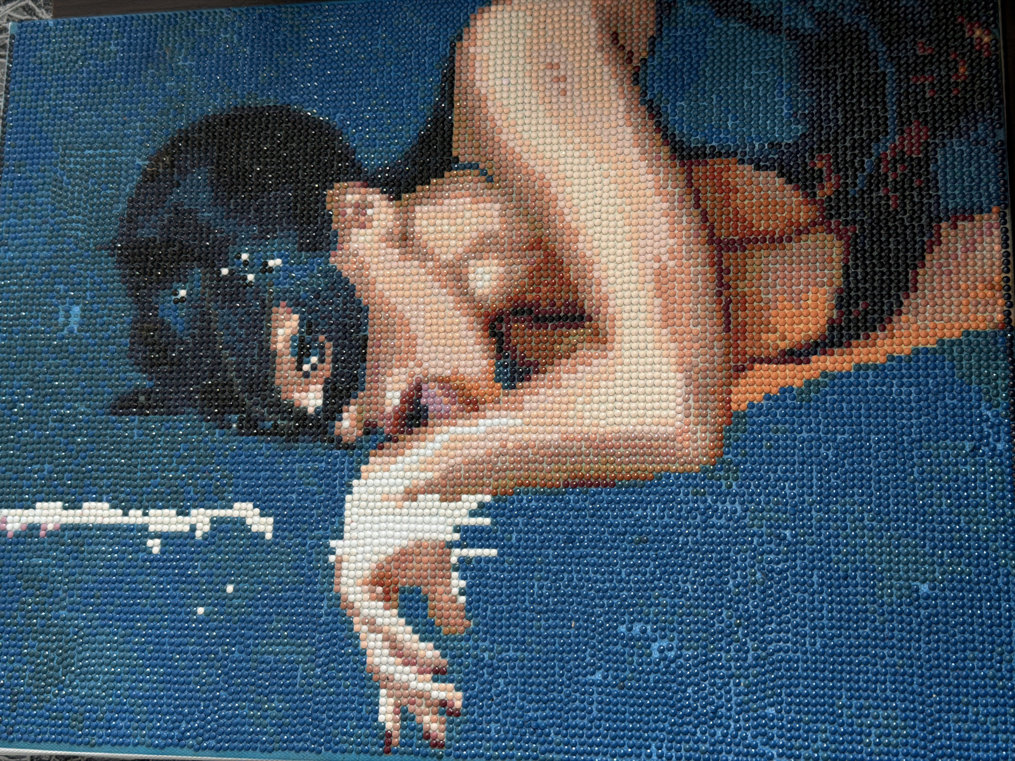 This is a detailed, close-up, eye-level shot of a diamond painting, depicting a woman in a black cat mask blowing white powder. The painting uses thousands of small, colored rhinestones to create the image. The background is a solid, dark blue, with some lighter blue speckles. The woman's skin tones are rendered with a variety of peach, pink, and light brown rhinestones. Her hair is black, and the mask is also black with white accents that appear to be stars or sparkles. Her right hand is extended forward, and she is blowing white powder from her mouth. Her left arm is bent, and her upper body is angled slightly away from the viewer. The overall impression is a dramatic and artistic representation. The location is not specified in the image.