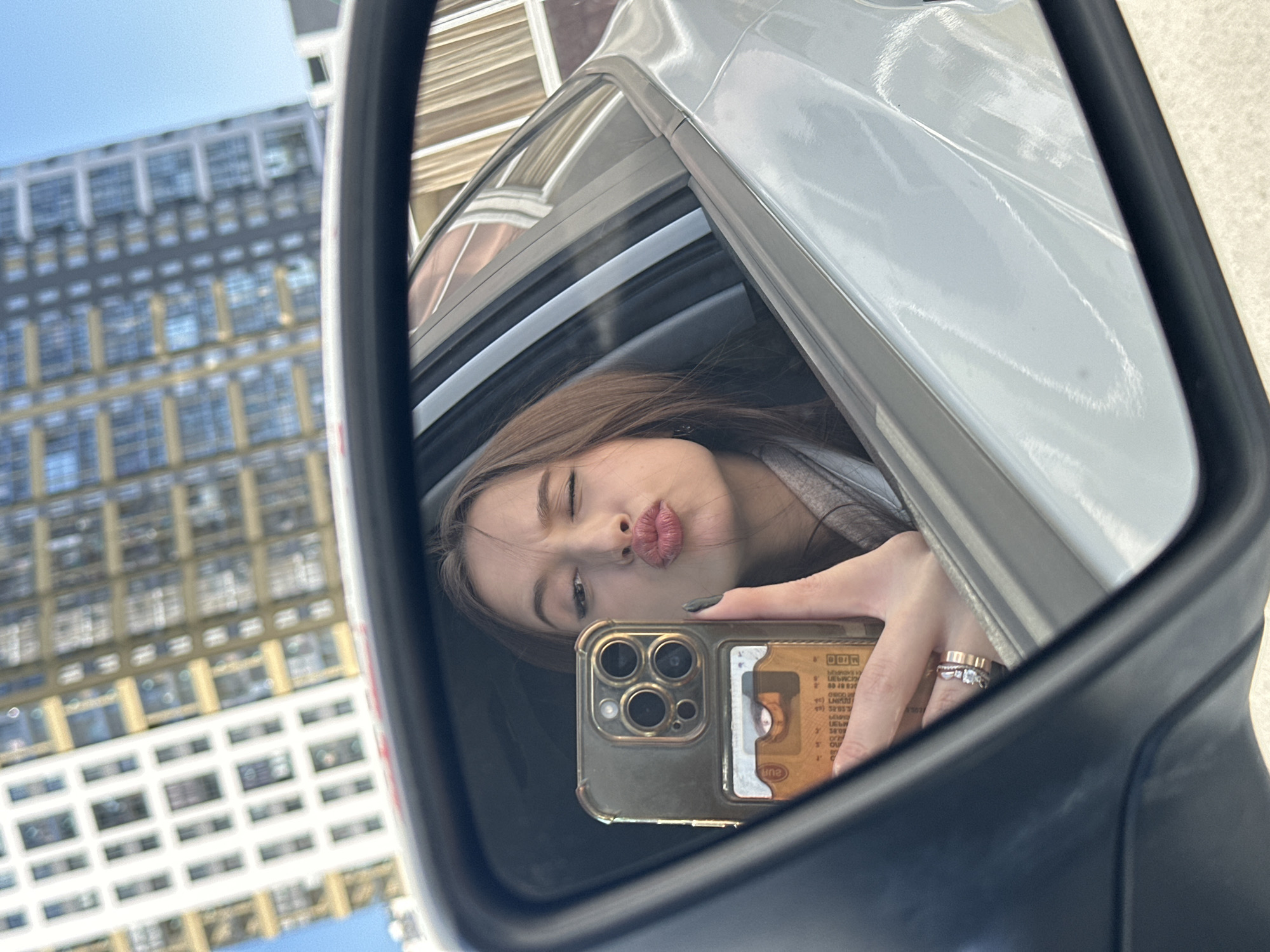 The image is a unique selfie captured in a car's side mirror in Yekaterinburg, Russia, during bright daytime. The central subject is a young woman, seen in reflection, posing with a playful "kiss face" expression. She has long brown hair and is looking towards the viewer. Her left hand is visible, holding a gold-colored smartphone with a transparent case. Several rings adorn her finger, including what appears to be a wedding or engagement band and another decorative ring.
Inside the phone's clear case, a card is visible, partially obscured, but displays the text "DON" and "EURO" along with other indistinct smaller characters. The reflection encompasses not only the woman and her phone but also a portion of the light-colored car's exterior (likely white or silver). In the background, beyond the car, several large, modern urban buildings with numerous windows are reflected in the mirror. The buildings are somewhat out of focus but clearly establish an urban setting under a clear, bright blue sky, indicating sunny weather. The image is oriented at an angle, characteristic of a casual mirror selfie, and conveys a lighthearted, confident mood.