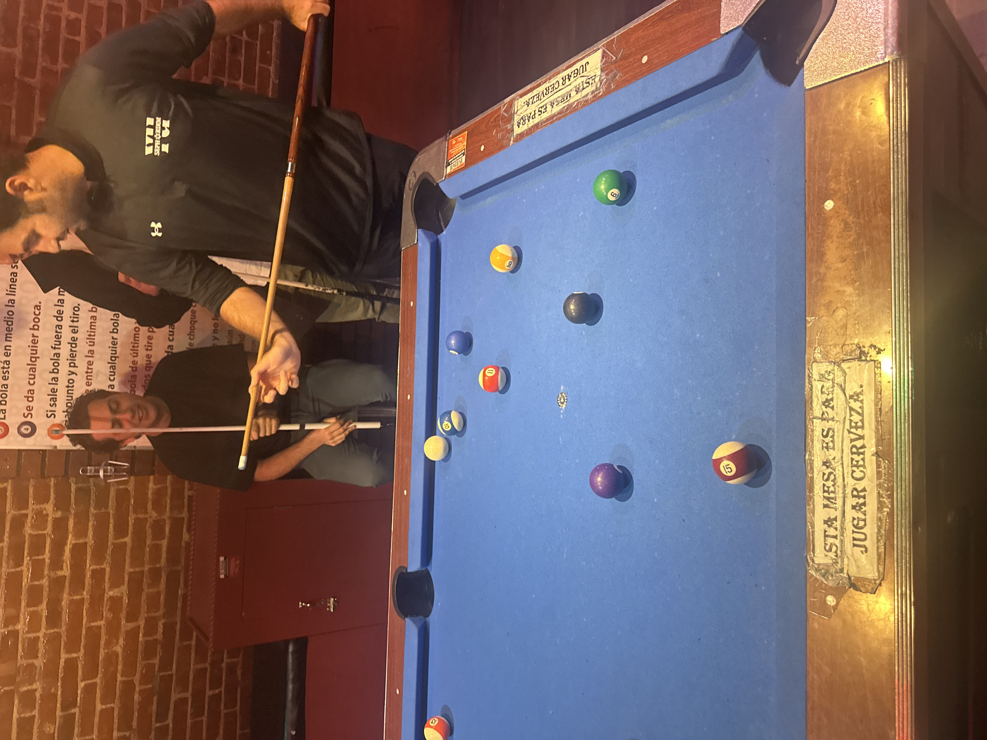 A medium shot depicts a billiard table with a blue felt surface, various numbered billiard balls scattered across it, and two men standing alongside it. The man closest to the camera holds a cue stick, seemingly preparing to strike the cue ball. In the background, a red brick wall is visible, with a sign written in Spanish. The billiard table has a worn appearance, with faded signage. The overall environment appears to be a dimly lit bar or entertainment establishment. The location is presumed to be outside of New York due to the Spanish sign.