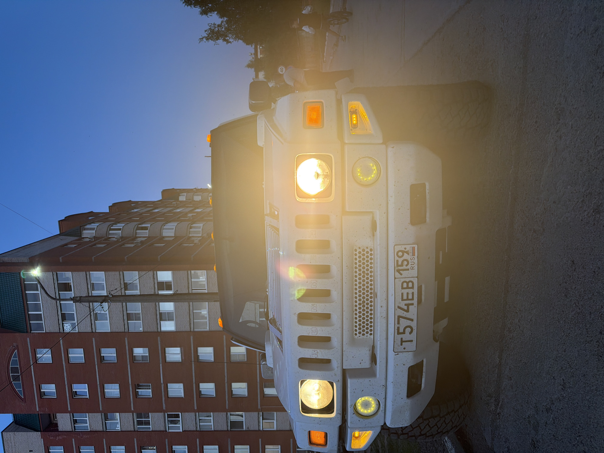 The image shows the front view of a white Hummer H2 SUV parked on an asphalt street in Chaikovsky, Russia. The vehicle's headlights are on, illuminating the surrounding area. The license plate reads "T574EB 159 RUS". Behind the vehicle is a multi-story brick building with multiple windows, reflecting the sky. Above the vehicle, the sky is visible, transitioning from blue to a lighter shade near the horizon. A street light pole is partially visible next to the building. At the top of the image, some greenery and a figure on a bicycle are visible. The overall lighting suggests it is dusk or dawn.