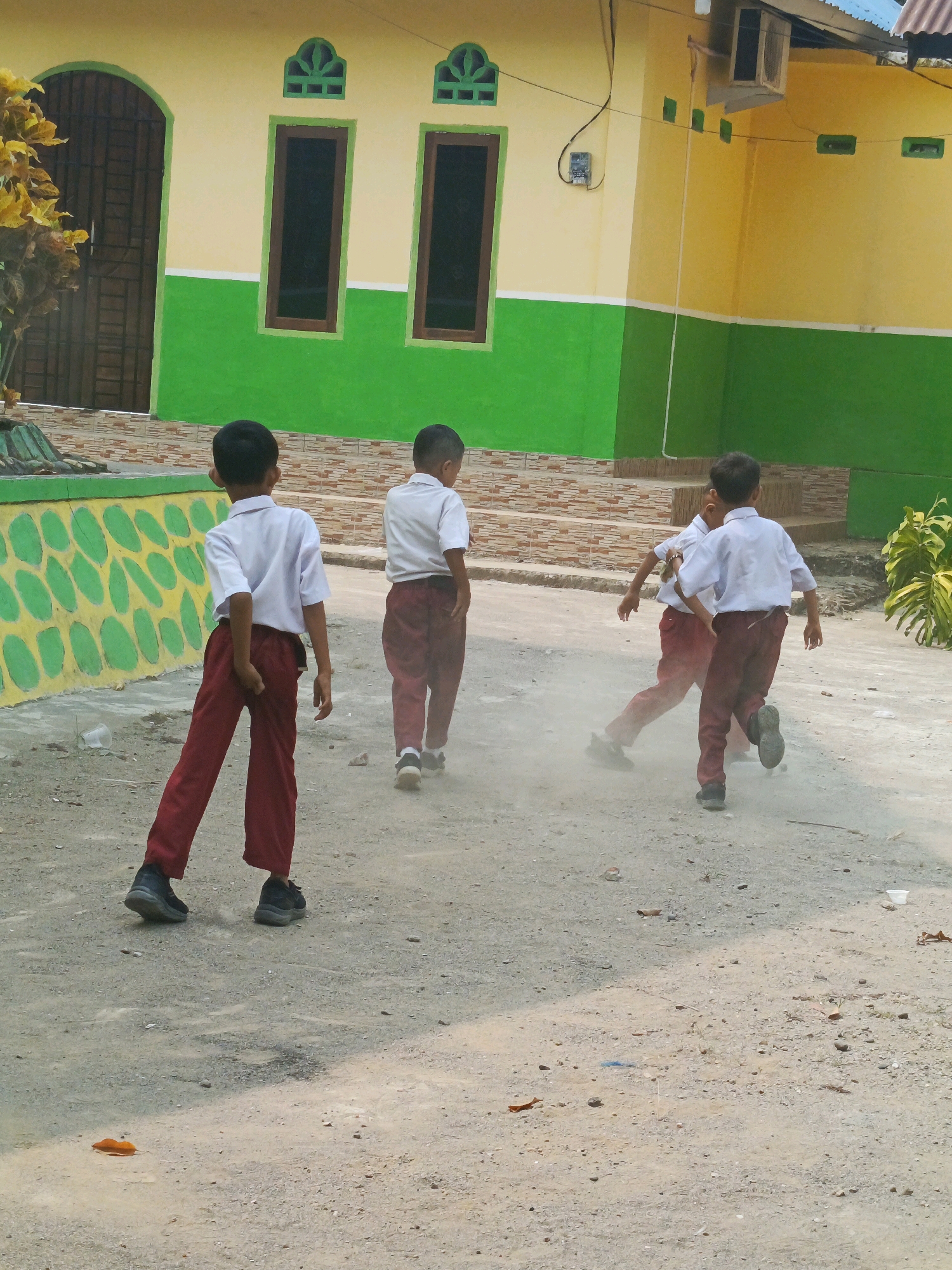 Four young boys, dressed in white shirts and dark red pants, are in a dusty outdoor area in front of a building. The building has a yellow upper half and a green lower half, with dark windows and decorative elements. The ground is a mixture of dirt and small stones, with some patches of light and shadow. One boy in the foreground is walking away from the camera, with his left hand near his back. Behind him, another boy is also walking away. Two other boys are in motion further ahead, with one kicking up dust. The scene appears to be on a school campus in Karya Dadi, Indonesia, possibly during a recess or outdoor activity.
