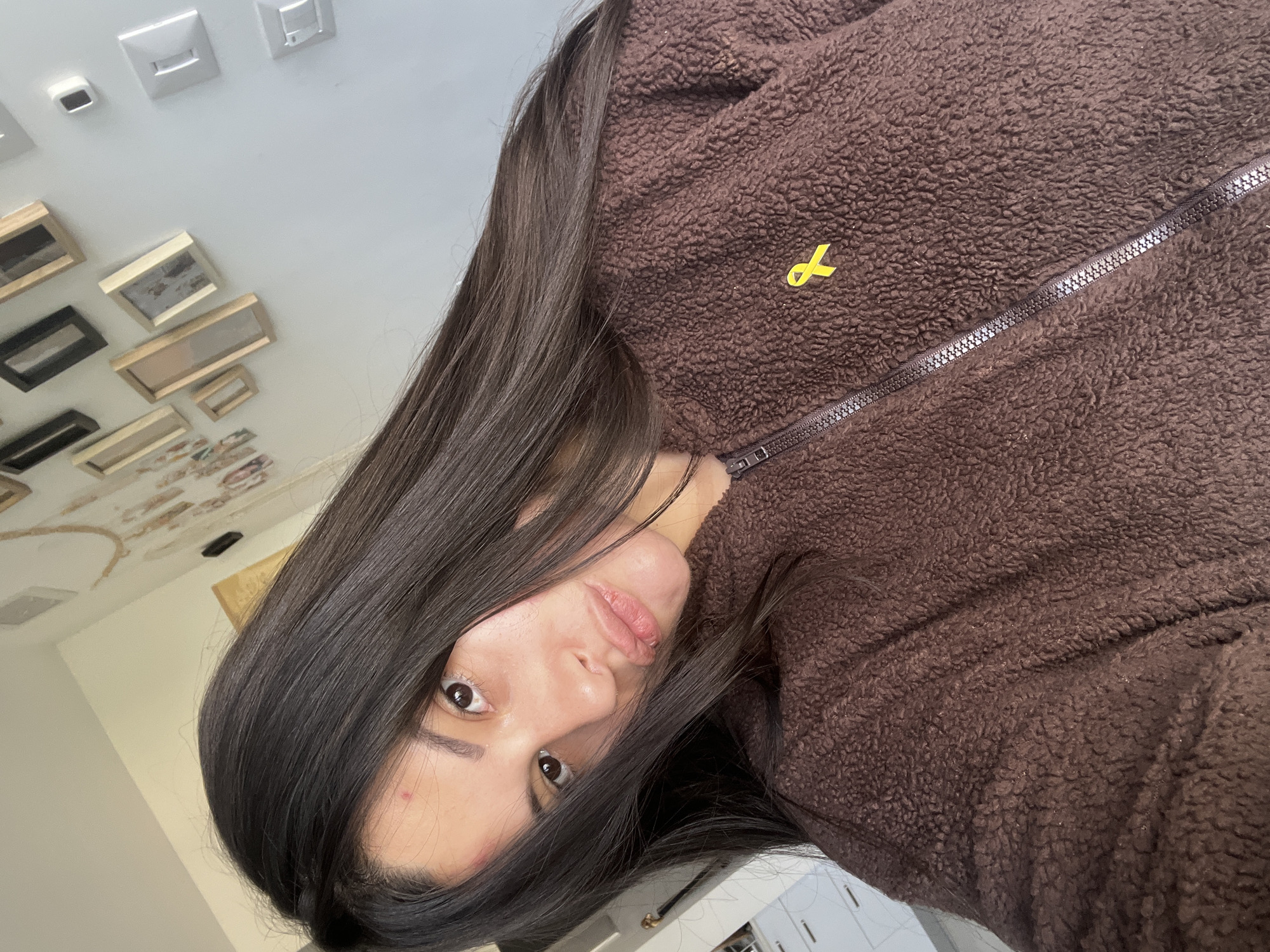 The image presents an angled, close-up view of a young woman, likely a selfie. She has long, straight dark hair that partially frames her face, and she looks directly at the camera with a neutral expression, her lips slightly parted in a soft pout. She wears a brown, textured fleece jacket with a dark zipper running diagonally across the front. A small, bright yellow ribbon pin, signifying a symbolic cause, is prominently attached to the left side of her jacket.
The background reveals an indoor setting with a light-colored ceiling. This ceiling is decorated with multiple white square and rectangular fixtures, possibly light switches or outlets, and an eclectic arrangement of various framed pictures and artworks of different sizes and orientations. The woman appears to be posing for the picture. The location context for this scene is Kiryat Bialik, Israel. There is no visible text in the image.