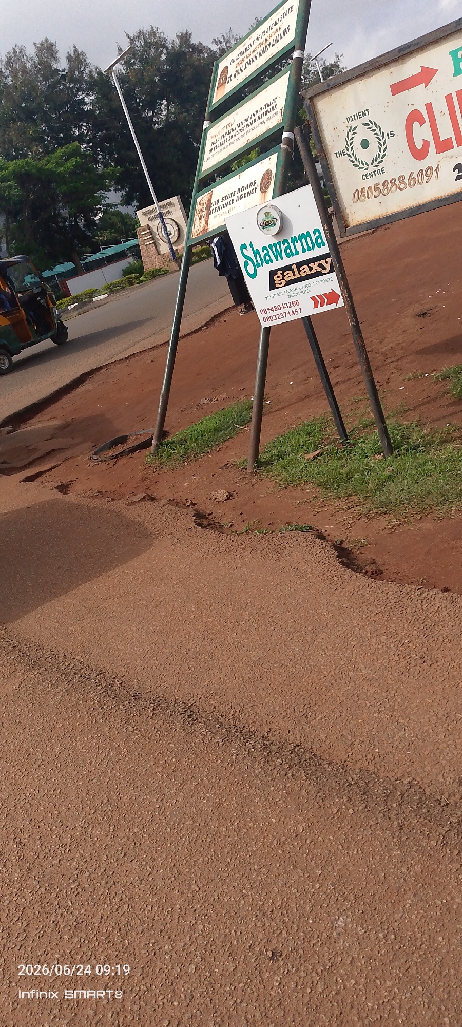 The image depicts a street scene in Jos, Nigeria. In the foreground, the rough, reddish-brown asphalt road surface is visible, with patches of grass and dirt along the side. A distinctive feature of the road is a dark, circular object embedded in the surface, possibly a manhole cover or drain. In the background, a white signpost stands tall, adorned with various green and white signs. One sign announces "Shawarma galaxy," with an arrow pointing to the right, and lists two phone numbers and an address: "9TH STREET, FEDERAL LOWCOST, OPPOSITE WILTON HOTEL." Another sign bears the text "GOVERNMENT OF PLATEAU STATE," followed by names and information about road projects. A third sign reads "PLATEAU STATE ROADS MAINTENANCE AGENCY." To the left of the signpost, a vibrant yellow and green auto-rickshaw, a common mode of transport in the region, is partially visible. The sky is overcast, suggesting a cloudy day. The timestamp "2026/06/24 09:19" and "Infinix SMARTS" are overlaid at the bottom left corner, indicating the date and time the photograph was taken and the device used. The overall scene conveys a sense of everyday life in a Nigerian city, with a focus on local businesses and government infrastructure.