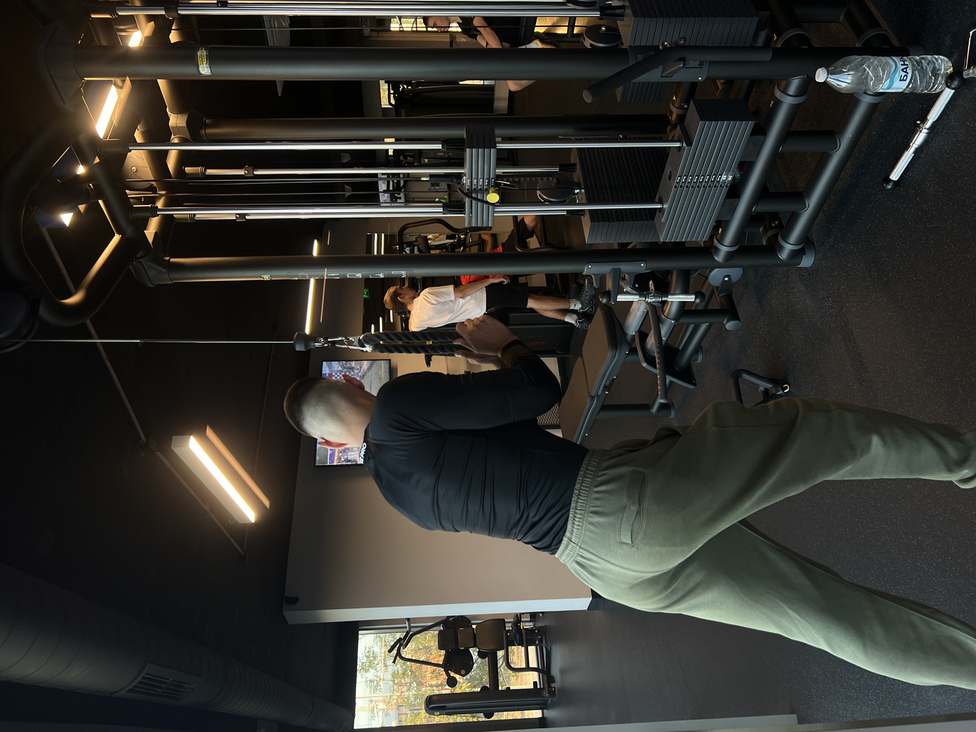 An eye-level, vertically oriented shot captures a man performing a cable exercise at a gym in Plovdiv, Bulgaria. The man, seen from the back, has a short haircut and is wearing a long-sleeved black shirt and olive-green sweatpants. He is gripping a rope attachment and engaging in an exercise, likely a triceps pushdown, on a large cable machine system.
The gym features dark walls, a black rubber floor, and bright, rectangular strip lighting on the ceiling. Various pieces of modern gym equipment are visible, including multiple weight stacks. One weight stack prominently displays numbers from "20" up to "200" in increments of "10." In the background, other gym-goers are present, with one man in a white t-shirt and another wearing glasses partially visible. A clear plastic water bottle with the brand "БАНКЯ" (Bankya) printed on it rests on a component of the gym machine. A yellow label on one of the background bars suggests the equipment brand "TECHNOGYM."
A television screen displaying what appears to be a sports or racing event is mounted on the wall. Large windows in the background reveal natural daylight and outdoor foliage, indicating it is daytime, though the interior is brightly lit by artificial lights. The scene depicts a functional and modern fitness environment.