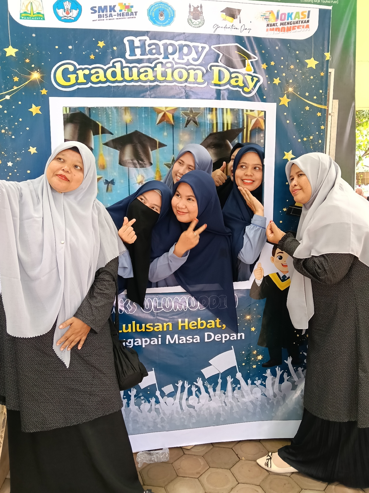 Five women are posing for a photo at a "Happy Graduation Day" photo booth. Two women wearing light grey hijabs and dark dresses stand on either side of the booth, while three women are posing within the booth's frame. One of the women within the booth is wearing a black niqab, and the other two women are wearing blue hijabs. The booth itself has a blue background with yellow stars and graduation caps. Text on the booth includes "Happy Graduation Day" and "Lulusan Hebat, gapai Masa Depan." Logos at the top of the booth identify the location as being related to SMK (Sekolah Menengah Kejuruan), a vocational high school, and also mentions "Cabang MQK Tauhid Putri," indicating a connection to religious education for women. A logo also states "LOKASI INDONESIA," a phrase which translates to "INDONESIA LOCATION" and "KUAT, MENGUATKAN INDONESIA" which translates to "STRONG, STRENGTHENING INDONESIA."