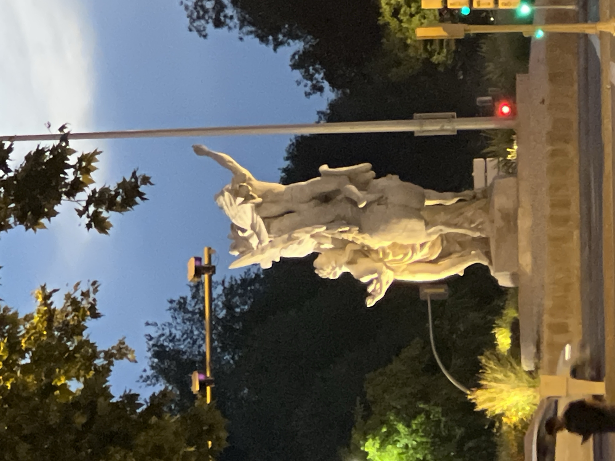 The image shows a white stone sculpture of three figures, likely a depiction of mythological characters, positioned atop a stone pedestal. The statue is partially illuminated by streetlights. Above and behind the statue are trees, with dark foliage against a sky transitioning from twilight blue to dark. A traffic light with green and red lights is visible above and to the right of the statue. A light pole with purple lights is to the left of the statue. The location is potentially Madrid, Spain.