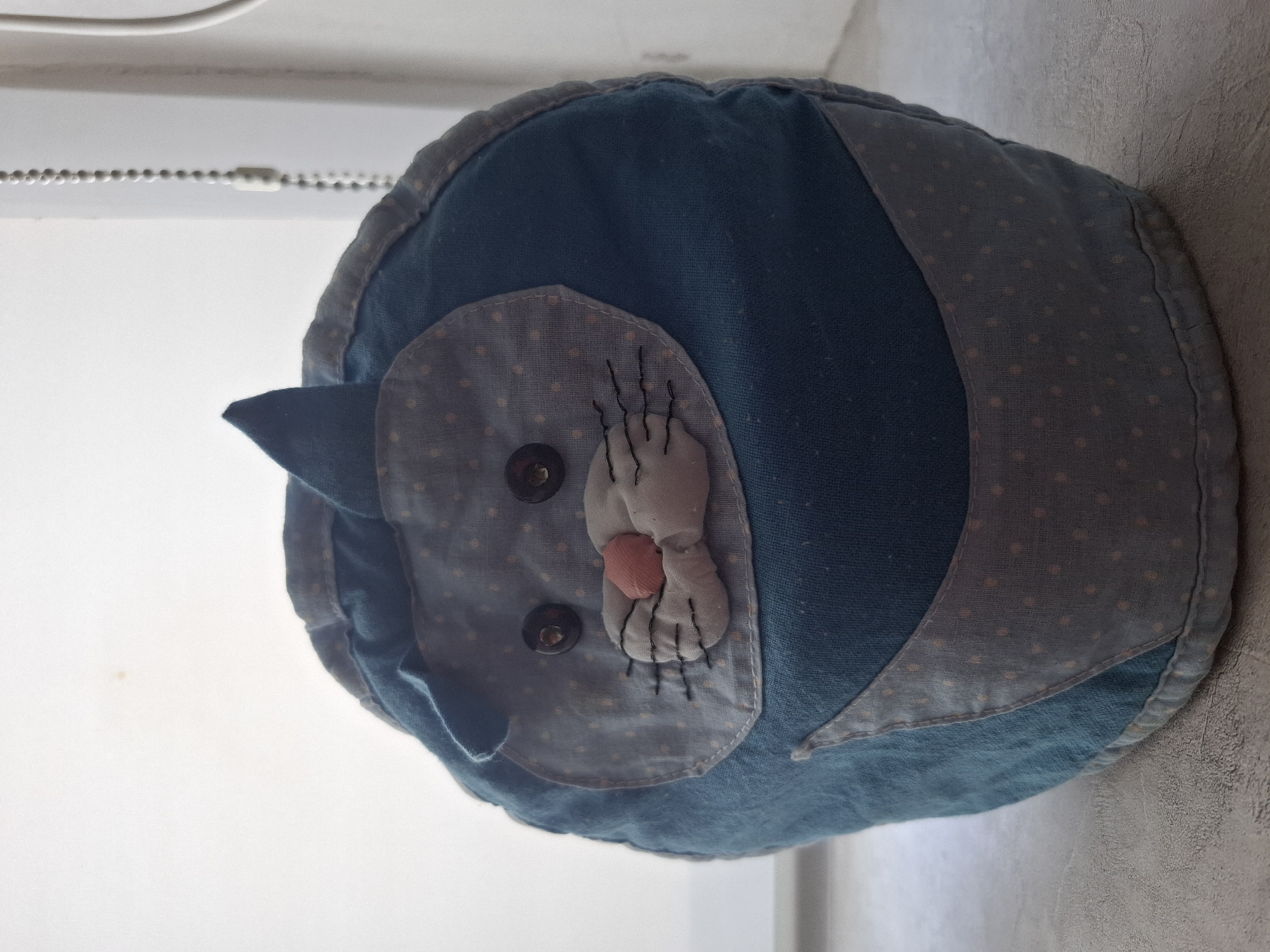 A handmade, blue cat-shaped pillow is positioned near a window. The pillow has a light blue, polka-dot fabric face with button eyes, a stitched-on white and pink nose, and sewn whiskers. Dark blue fabric forms the body of the cat. The pillow appears to be resting on a light-colored surface, possibly a windowsill. The scene's location is in Амзя, Russia.