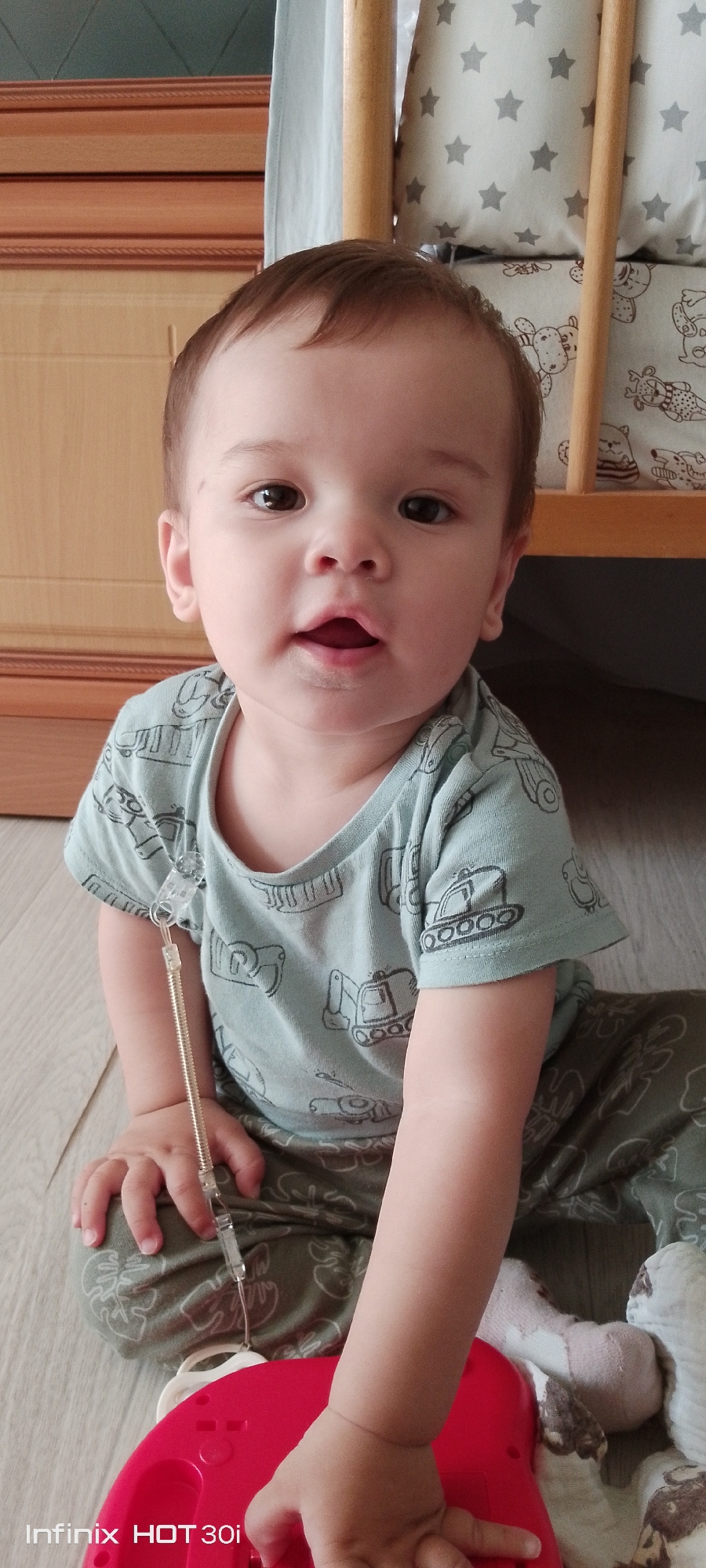 A young child, appearing to be around one to two years old, is sitting on a wooden floor. The child has fair skin, brown eyes, and short brown hair. They are wearing a light blue t-shirt with a pattern of construction vehicles and dark green pants with a leaf pattern. A clear plastic cord with a coiled spring is attached to the child's t-shirt, extending downwards towards an unseen object. The child is looking directly at the camera with an open mouth. In the background, a wooden crib with patterned bedding is visible, along with a section of a wooden cabinet. The lighting suggests an indoor setting, likely a home. The image is watermarked with "Infinix HOT 30i" in the lower left corner. The specific location of Kedrovy, Russia, cannot be determined from the visual information provided in the image.