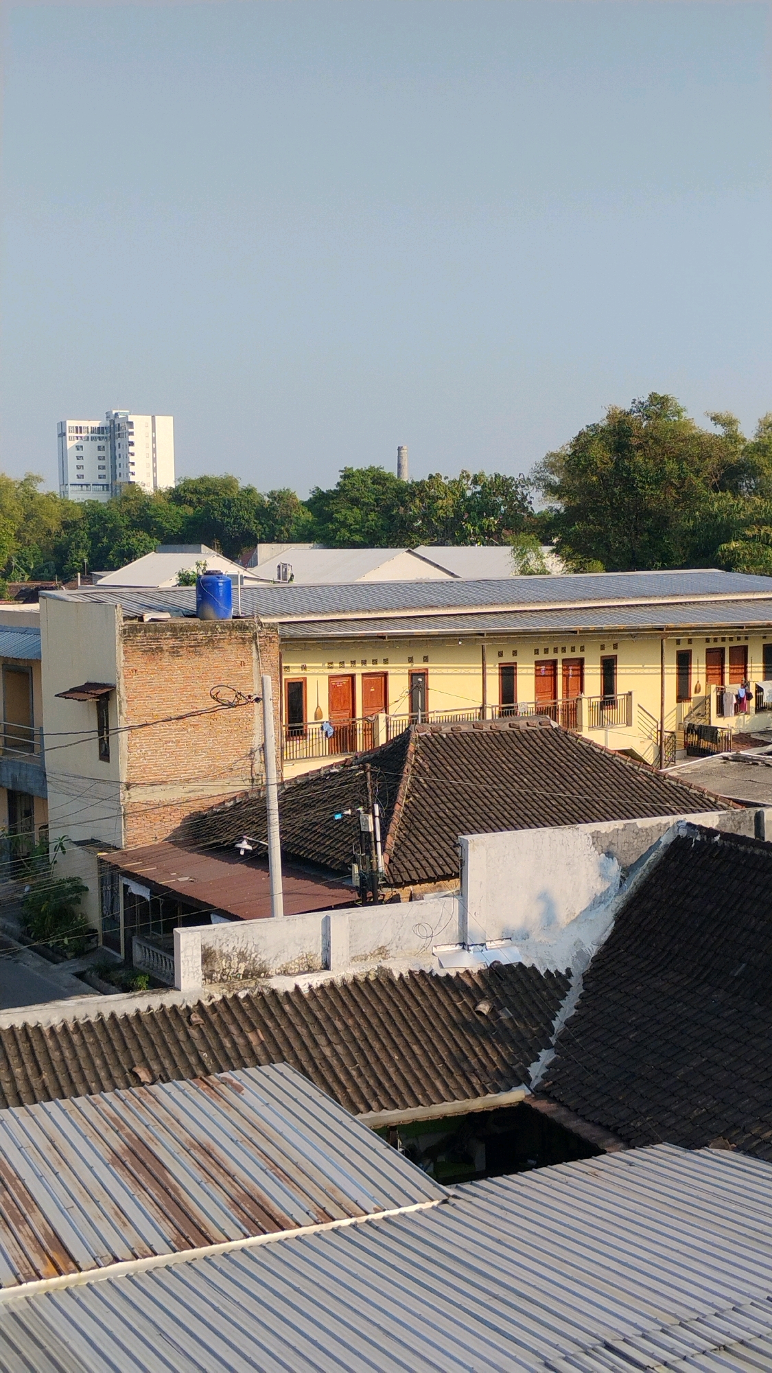 The image depicts a view of rooftops in Gumpang, Indonesia, under a clear, pale blue sky. In the background, a modern, multi-story white building stands tall, contrasting with the surrounding trees and a cylindrical industrial structure or chimney. The foreground and mid-ground are dominated by a variety of roof structures. One prominent building features a yellow facade with multiple dark wooden doors and windows, accessible by balconies with railings. This building also has a brick section on one side topped with a blue water tank. Several tiled roofs, varying in color from dark gray to brown, are visible, some showing signs of age. In the foreground, corrugated metal roofs are prominent, one with visible rust streaks. A utility pole with electrical wires runs vertically through the scene. The overall impression is an urban or semi-urban residential or commercial area.