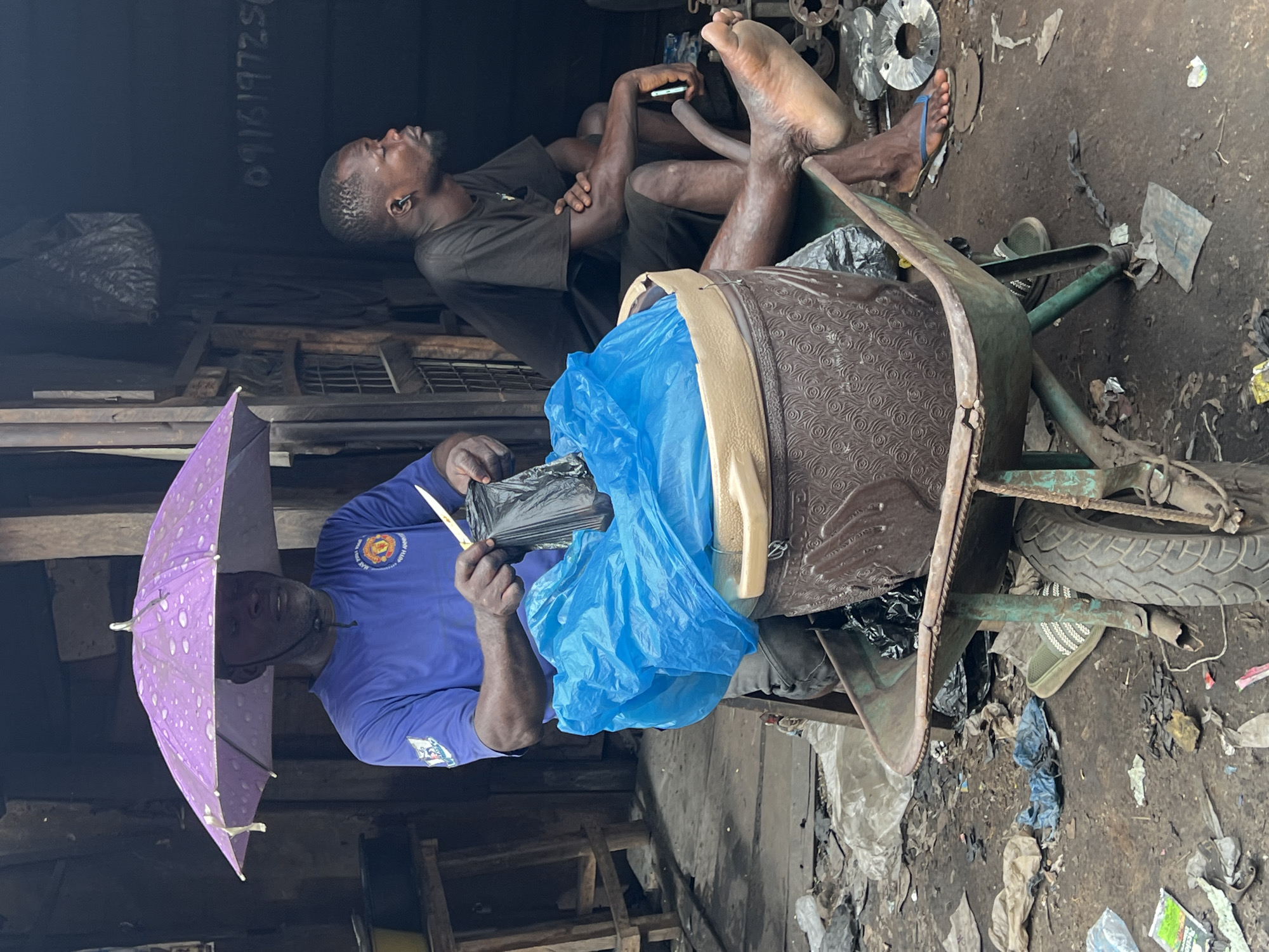 The image depicts a scene in Nkpor, Nigeria. A man stands under a purple umbrella, holding a knife and a black plastic bag. He stands next to a wheelbarrow, which contains a brown patterned suitcase covered with a blue plastic bag. Another man sits in the wheelbarrow with his bare feet propped up on a pile of metal objects. The man in the wheelbarrow is holding a cell phone. The environment appears cluttered, with litter and debris scattered on the ground. The scene is set inside or near a metal shop.