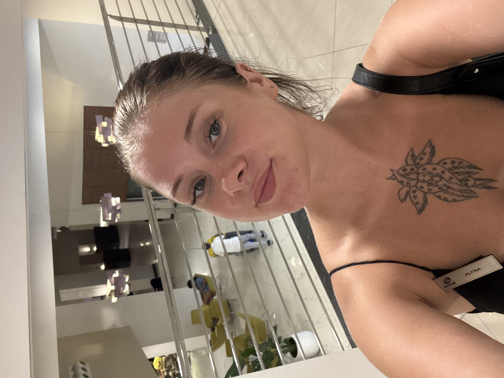 A young woman with light skin, brown hair pulled back, and blue eyes takes a selfie in a hotel lobby. She is wearing a black dress with thin straps and has a tattoo on her chest. A name tag is visible on her left strap, reading "Limak ALENA". The background shows a multi-level hotel interior with yellow chairs, potted plants, and staircases with metal railings. Two men, one in a white shirt and blue pants and another in a blue shirt, are visible in the background. Modern light fixtures hang from the ceiling. The overall scene suggests a hotel in Aksu, Turkey.