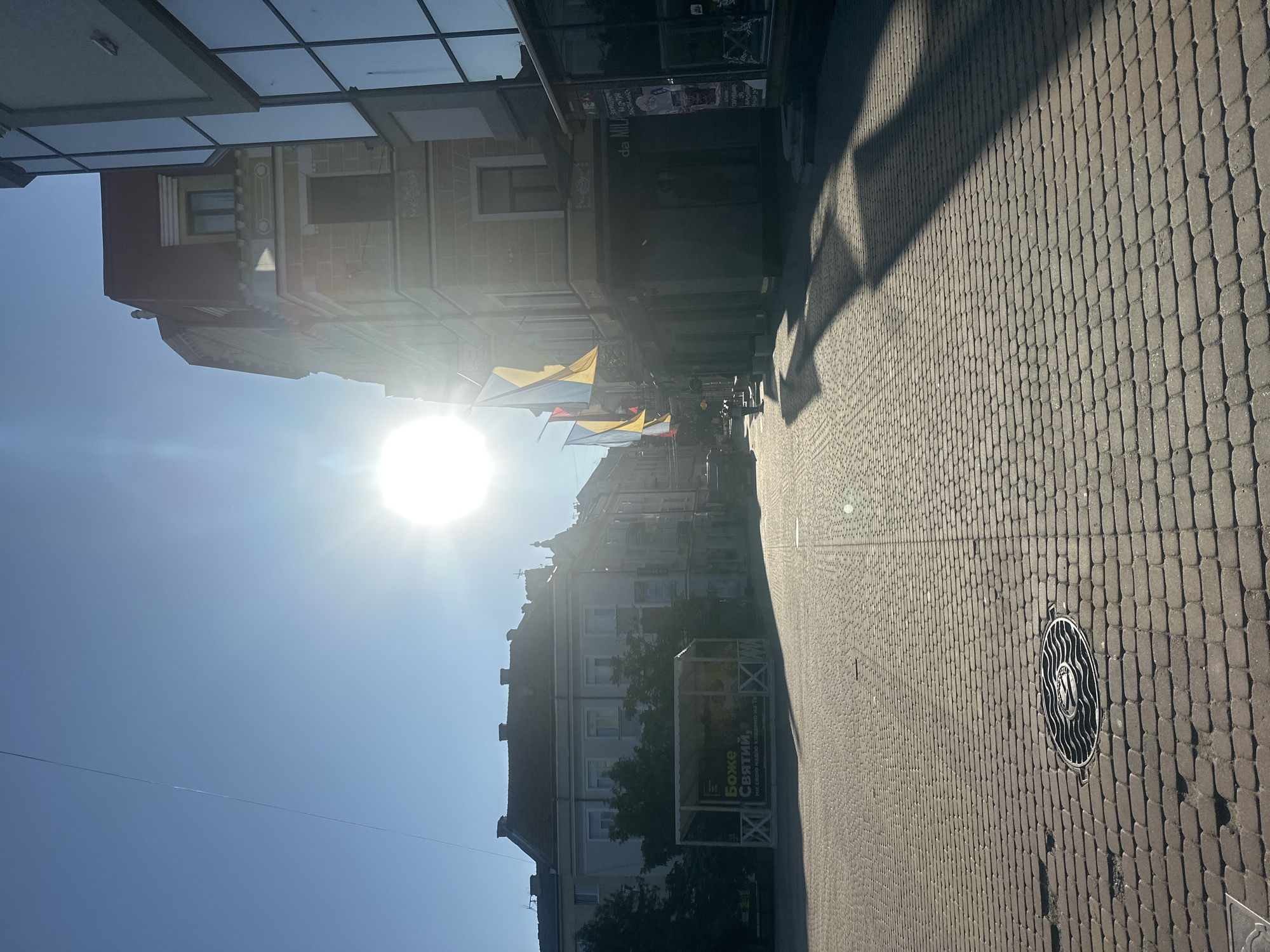 The image shows a view of Teatralna Square in Ternopil, Ukraine, on a sunny day. The sun is bright and high in the sky, casting long shadows across the cobblestone pavement. Several buildings line the square, with architectural styles varying from older, ornate structures to more modern glass-fronted buildings. Ukrainian flags are visible, hanging from some of the buildings. In the foreground, the cobblestone pavement is detailed with a manhole cover. A white gazebo with a sign is visible on the left side of the square. The scene depicts a quiet urban environment with no people immediately apparent.