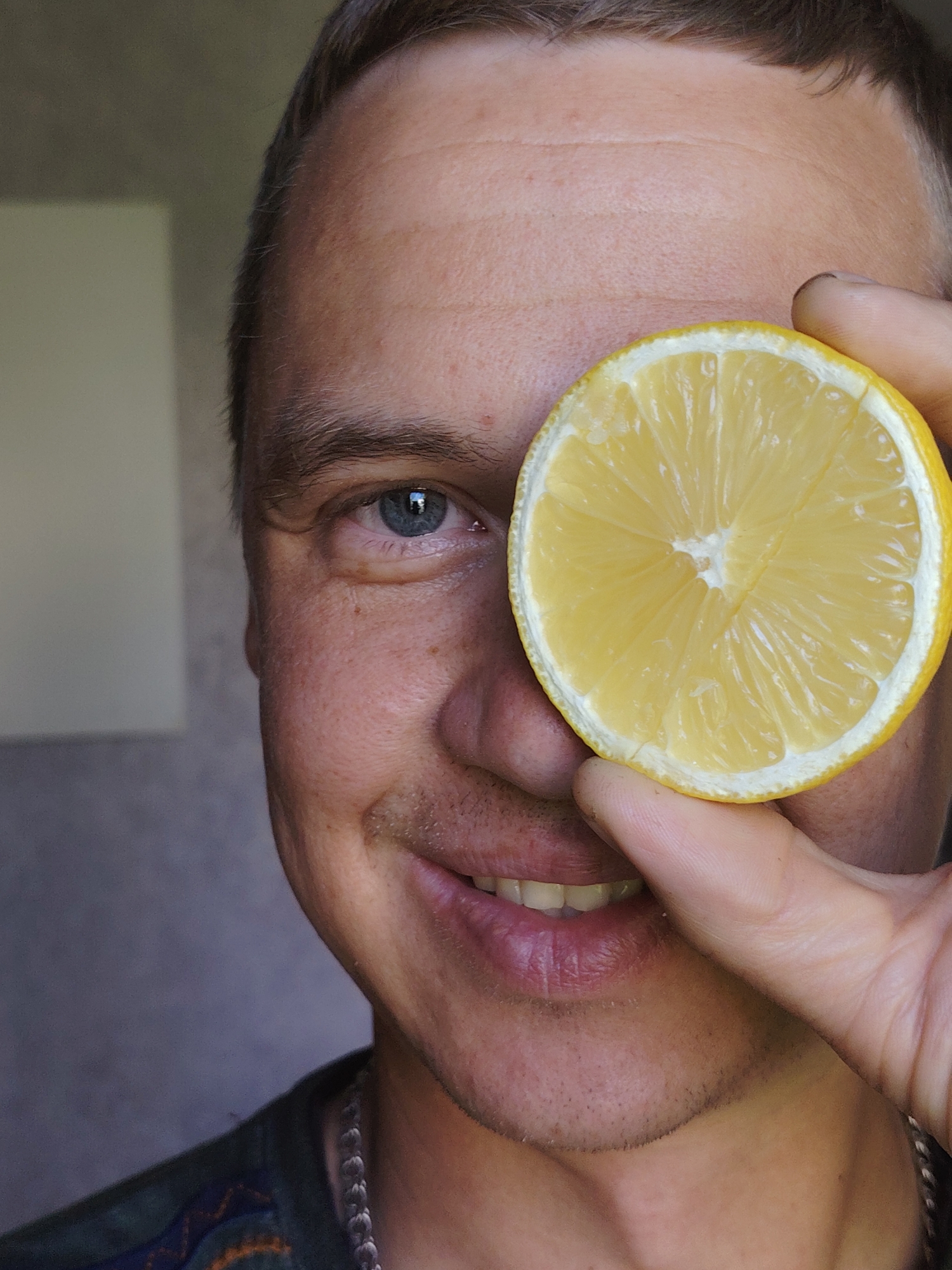 A close-up shot shows a man holding a half-cut lemon in front of his face, obscuring one eye. The man has short brown hair, fair skin, and blue eyes. He is smiling slightly, revealing his teeth. He is wearing a dark-colored shirt with a patterned design and a silver chain around his neck. The background is out of focus, showing a neutral-colored wall with a rectangular white object. The image appears to have been taken indoors with natural lighting. There is no information available in the image that would indicate that the location is in the city of Chaikovsky, Russia.
