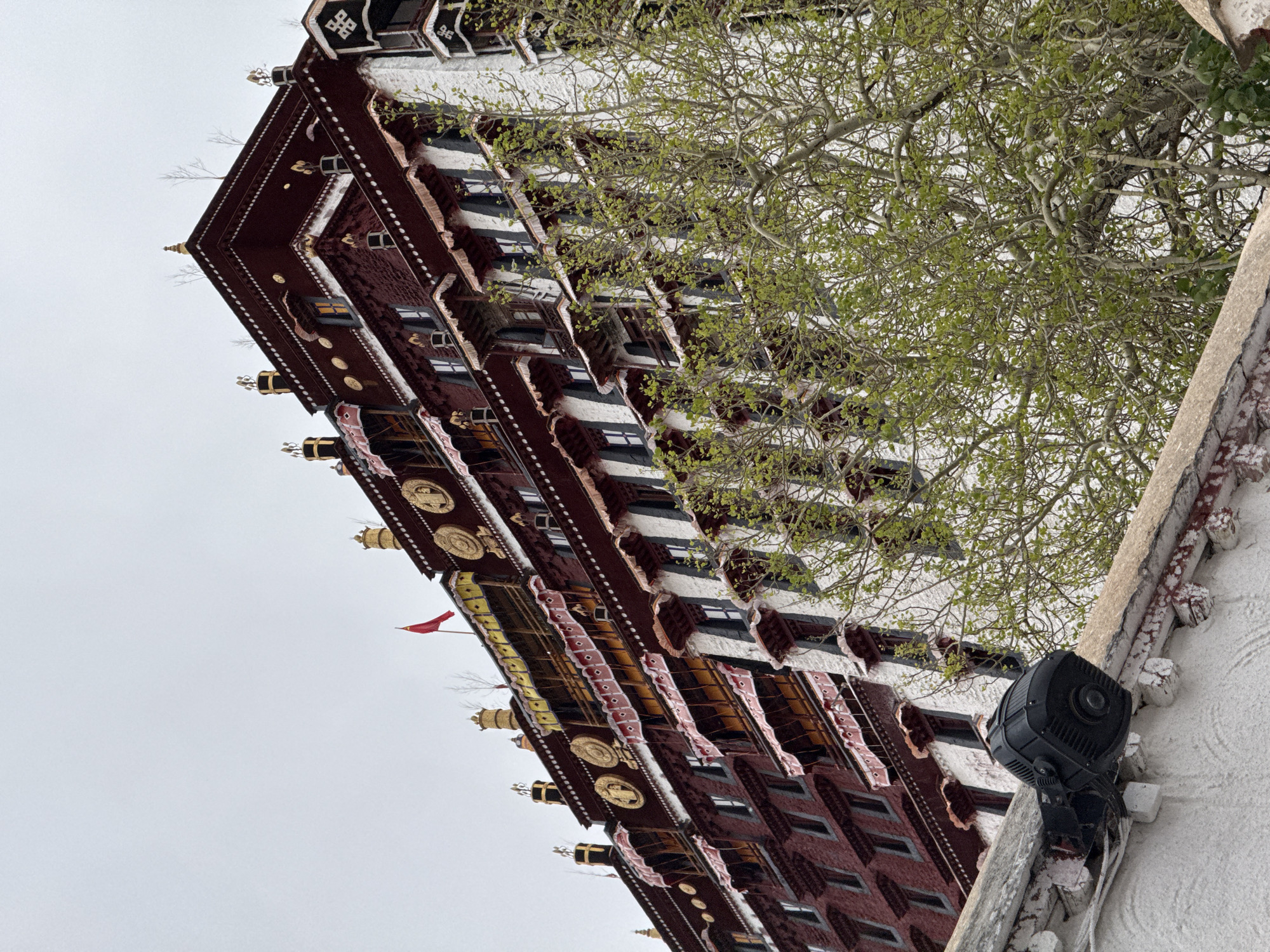 The image presents an upward-angled view of a traditional Tibetan building facade in Chengguan District, Lhasa, China. The building's exterior is characterized by a combination of dark red brickwork, white decorative elements, and black-and-white detailing on the upper sections. Windows are visible, alternating with the architectural elements. A sparse tree with budding green leaves is seen partially obscuring the building. A red flag flutters from the building. A black security camera is mounted on the building's white wall. The sky is overcast and light gray.