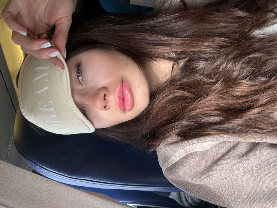 A young adult female is depicted in the cabin of an aircraft, likely within the vicinity of Lyon-Saint Exupéry Airport in Colombier-Saugnieu, France. She has long, wavy dark brown hair and fair skin. Her blue eyes are visible, and her lips are full with a glossy pink finish. She is wearing a light brown or beige ribbed sweater. Her right hand, featuring white manicured fingernails and a golden ring on the ring finger, is holding a light beige eye mask with the word "DREAM" printed on it, lifted to expose her right eye. Behind her, a dark blue aircraft seat is visible, confirming the interior setting of an airplane cabin. The left side of the frame shows a portion of the aircraft's interior wall or window structure. The subject is looking directly towards the camera.