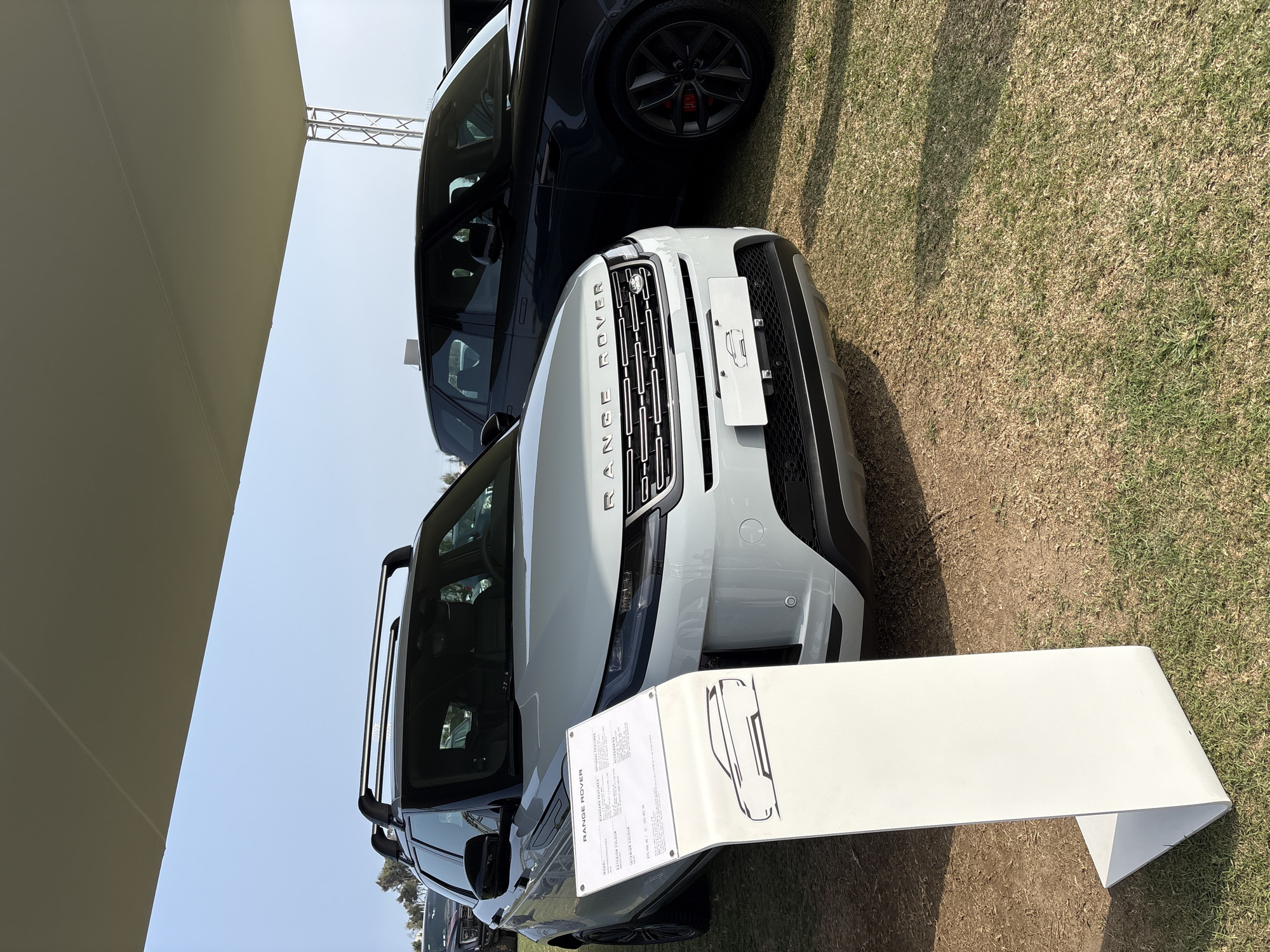 The image shows two Range Rover vehicles parked on a grassy area under a large canopy. The car in the foreground is a light-colored SUV, possibly white or silver, with a roof rack. In front of it, a white display stand with text and a sketch of an SUV is positioned. The Range Rover lettering is visible on the front grille. A dark-colored SUV, also a Range Rover, is parked to the right of the foreground vehicle. The environment appears to be outdoors, with dry grass and dirt on the ground. The sky is visible above the canopy. The scene suggests a car exhibition or dealership event. There are no people clearly visible in the frame. The location is described as the city of Lurín, Peru.