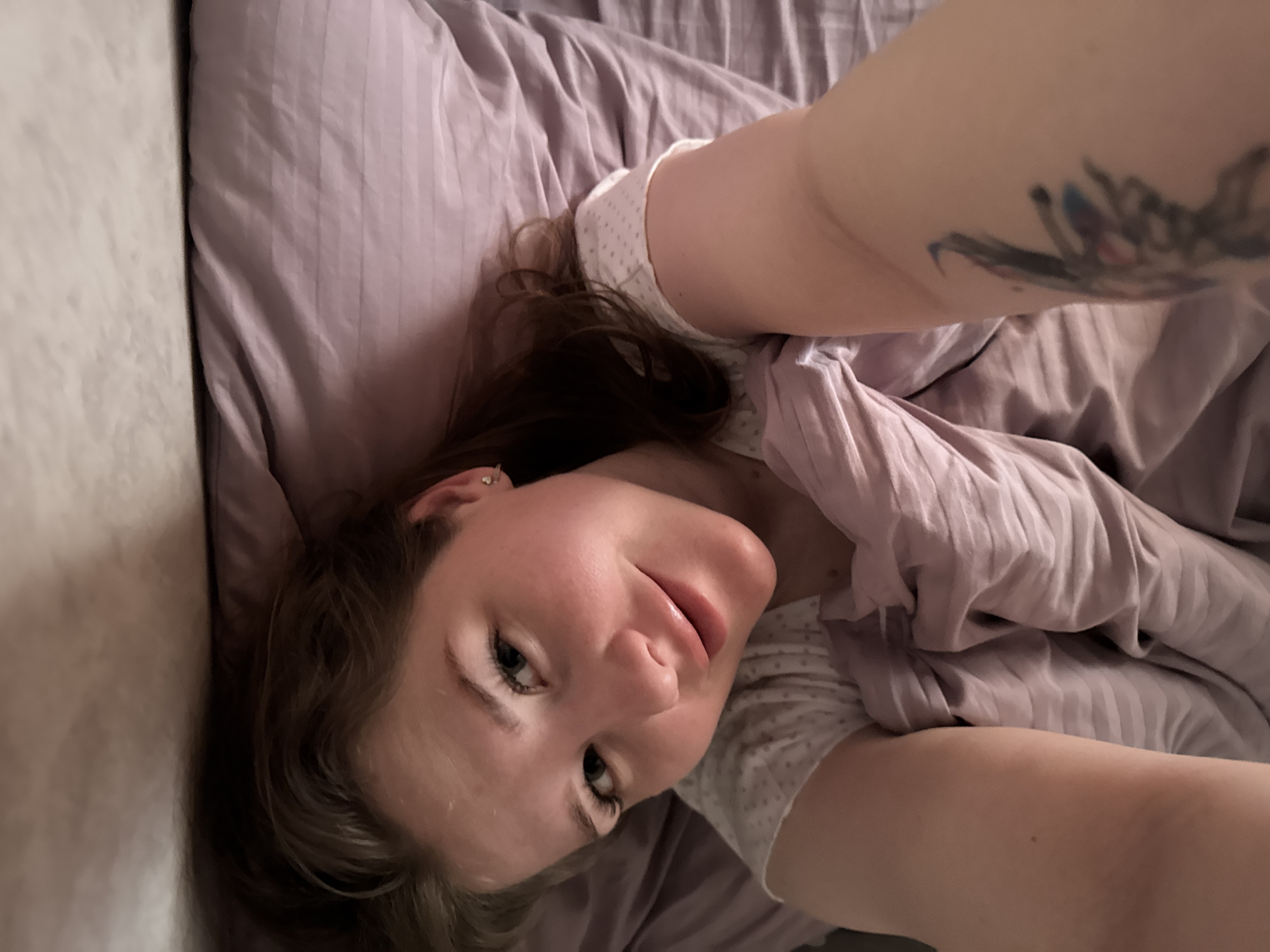 A woman is lying on a bed covered with lavender-colored sheets and a comforter. She is wearing a white shirt with small polka dots. She has brown hair and fair skin. A tattoo is visible on her arm. The headboard of the bed is visible in the background. The image was likely taken indoors. The city is not possible to ascertain from the image.