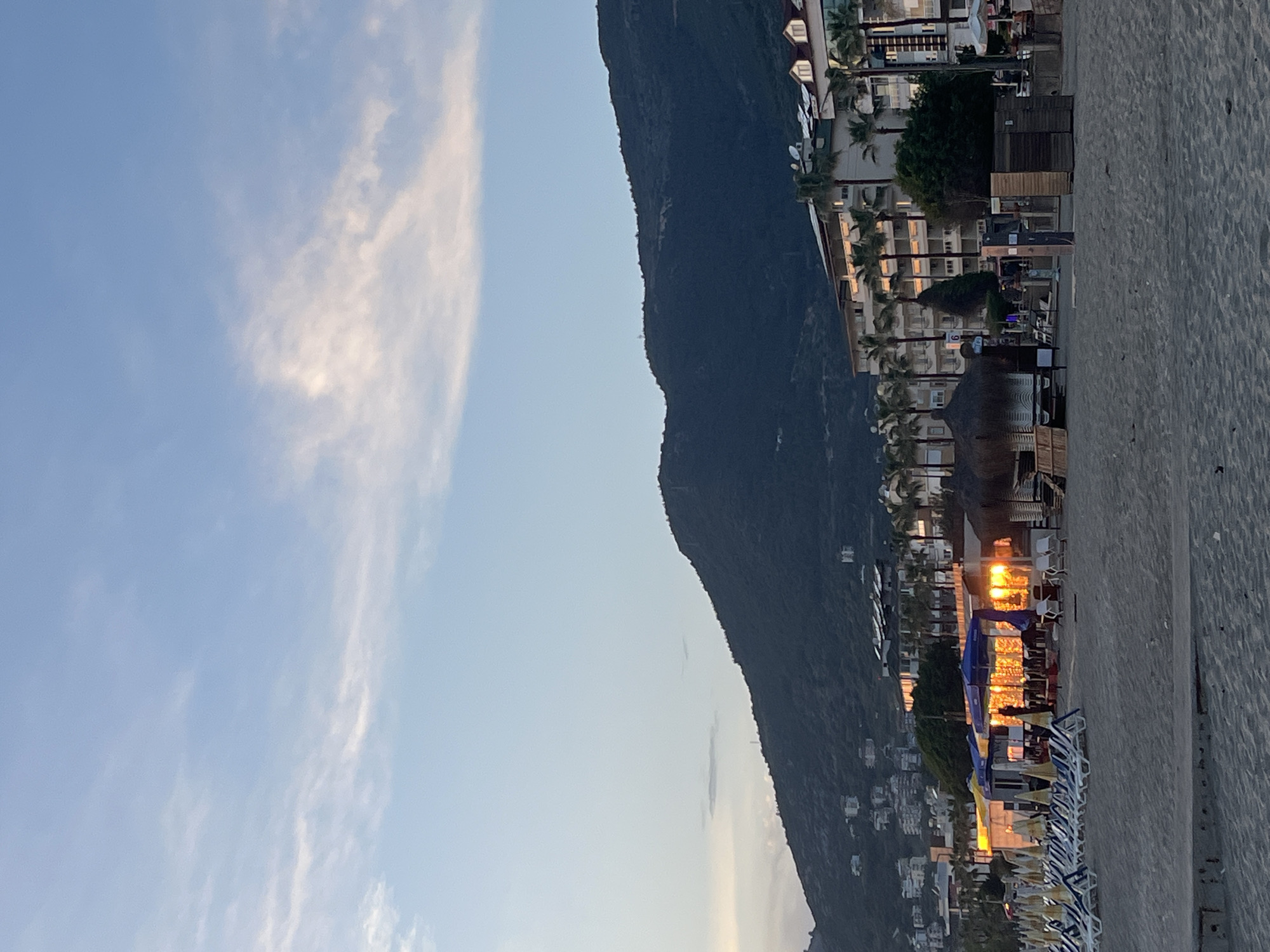 The image depicts a tranquil coastal scene in Alanya, Turkey, captured during either sunrise or sunset, indicated by the warm, golden light. A vast, dark mountain range dominates the left side of the frame, its silhouette rising against a light blue sky dotted with soft, wispy white clouds.
Along the mountain's base, a series of multi-story buildings, likely resort hotels, line the coast. These structures feature balconies and are brightly illuminated by the low sun, which casts a golden glow and creates prominent reflections on their facades. Numerous palm trees are interspersed among the buildings, enhancing the resort-like ambiance.
In the foreground, a wide, sandy beach stretches across the scene. Rows of empty beach loungers, many with blue and white striped cushions, are neatly arranged on the sand. A few straw-roofed structures, possibly umbrellas or small kiosks, are also visible on the beach.
No human activity is depicted, contributing to the serene and peaceful atmosphere. The golden hour lighting beautifully highlights the contrast between the dark mountain and the illuminated buildings and beach. No discernible text is visible in the image.