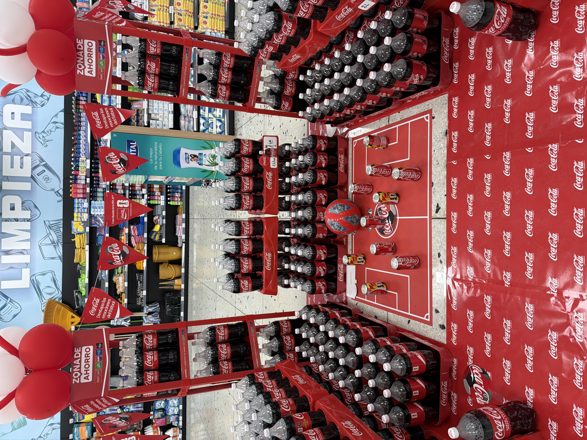 The image depicts a promotional display of Coca-Cola products inside a supermarket, likely in Turmero, Venezuela, given the context of a previous query about this location. The display is arranged with multiple shelves of Coca-Cola bottles and cans. A central element is a red display stand featuring Coca-Cola bottles, topped with red and white balloons. Several red triangular flags with the Coca-Cola logo are also visible, adding to the festive atmosphere. A miniature soccer field graphic is present on the floor, with cans and a soccer ball on it, suggesting a tie-in with a football event, possibly the World Cup. The overall environment is brightly lit, typical of a retail setting, with shelves of other supermarket products visible in the background. There are no people present in the image.