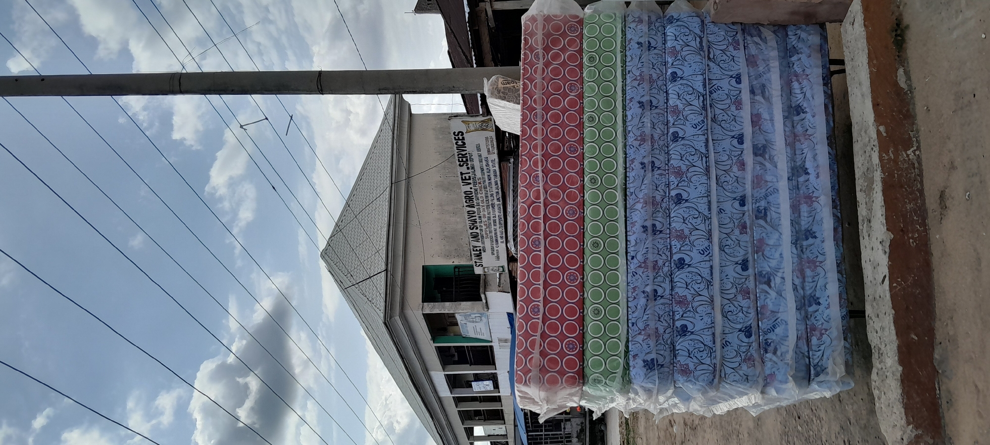 The image depicts a stack of mattresses covered in plastic packaging, situated outdoors on an unpaved surface. The mattresses have different patterned covers: the top one is red with a circular design, the second is green with a similar circular design, and the bottom three are blue with floral patterns. Behind the mattresses, a light-colored building with a grey, tiled roof is visible. A sign on the building reads "STANLEY AND SHAYO AGRO. VET. SERVICES" and provides a contact number and address in Jalingo, Taraba State, Nigeria. A concrete curb is in the foreground. Utility poles and power lines traverse the sky, which is partly cloudy. The scene appears to be in an urban or semi-urban area.