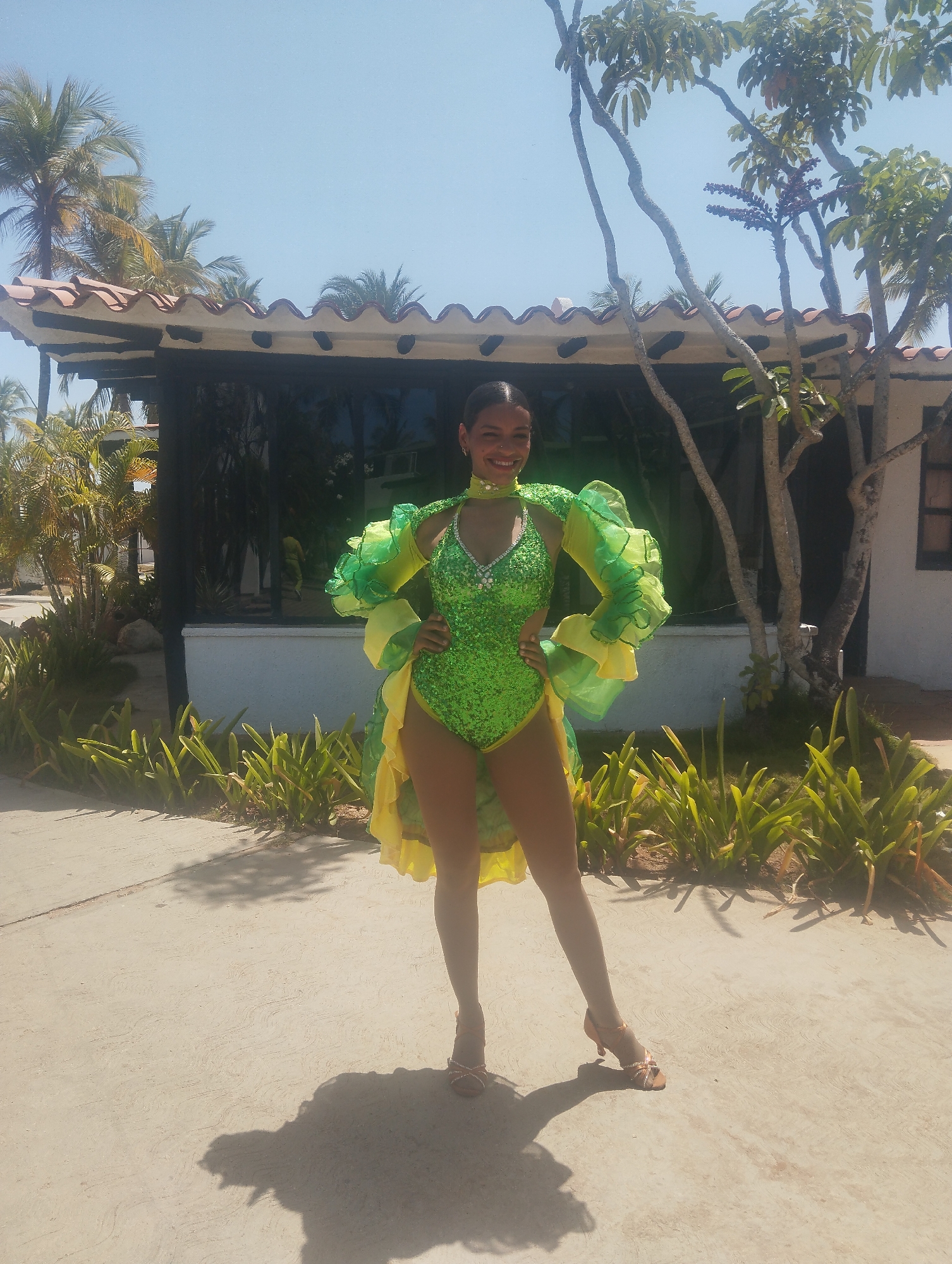 A woman is standing outside in Porlamar, Venezuela. The location appears to be a resort. She is wearing a bright green, sequined bodysuit with a yellow and green ruffled shawl and high heels. She is posing for the camera. In the background, there are palm trees, a building with black windows and a red tile roof, and landscaping.