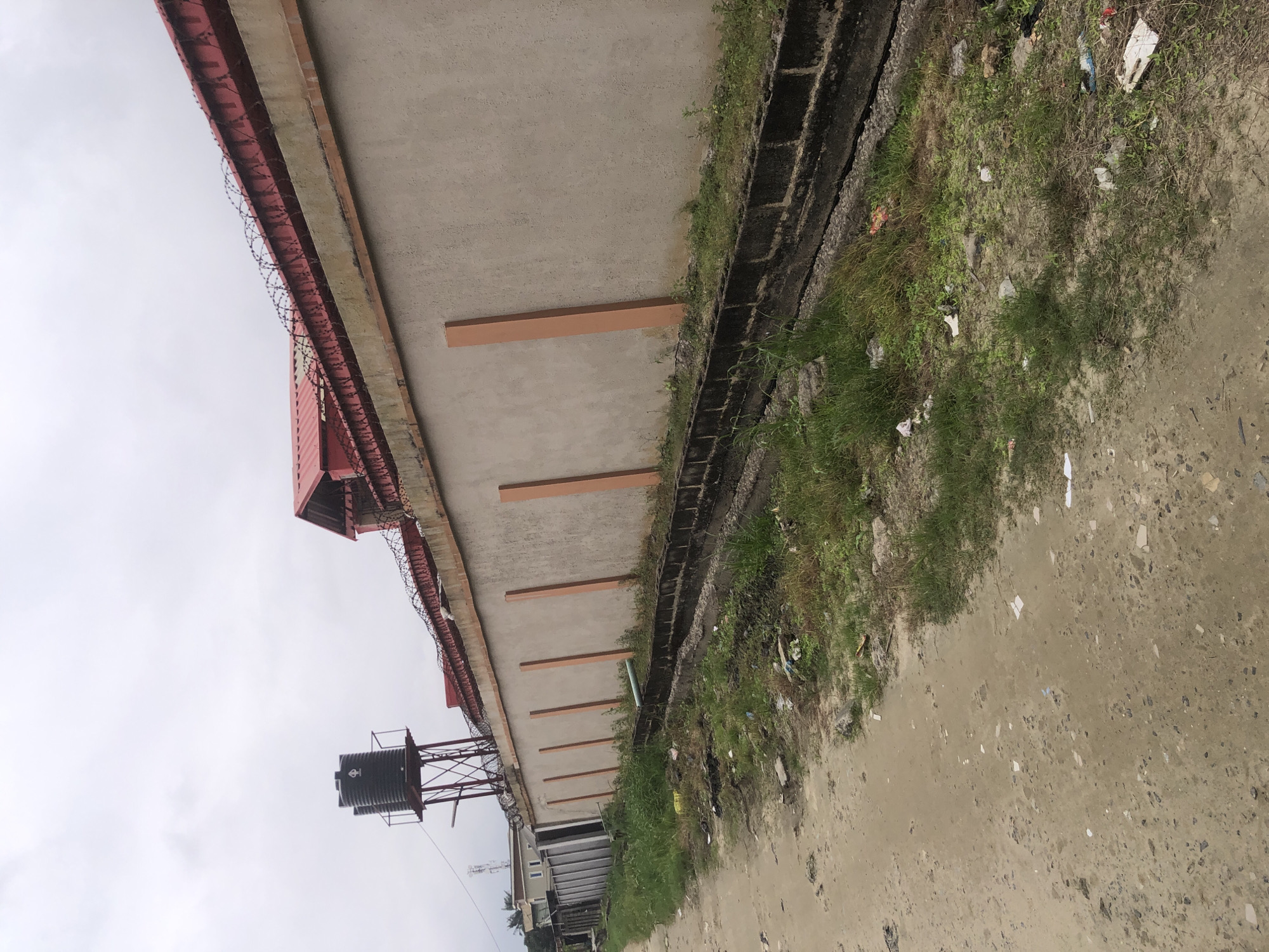 The image shows a street view in Ado, Nigeria, on an overcast day. A tall, grey concrete wall with an orange-tinted, sloped concrete cap runs along the right side of the frame. Barbed wire is attached to the top of the cap, and red metal roofing is visible behind it. On top of the wall, towards the left, a black water tank on a red metal support structure is visible. The wall is punctuated by regularly spaced, vertical, orange-tinted rectangular elements. A narrow, dark, concrete drainage ditch runs along the base of the wall, filled with some debris and overgrown with grass. To the left of the wall and ditch, a dirt road or path with scattered litter and sparse vegetation is present. Further left, a gray metal fence and a building with light-colored walls and dark windows are visible in the distance. The overall impression is of an urban environment with signs of wear and tear, including litter and overgrown areas.