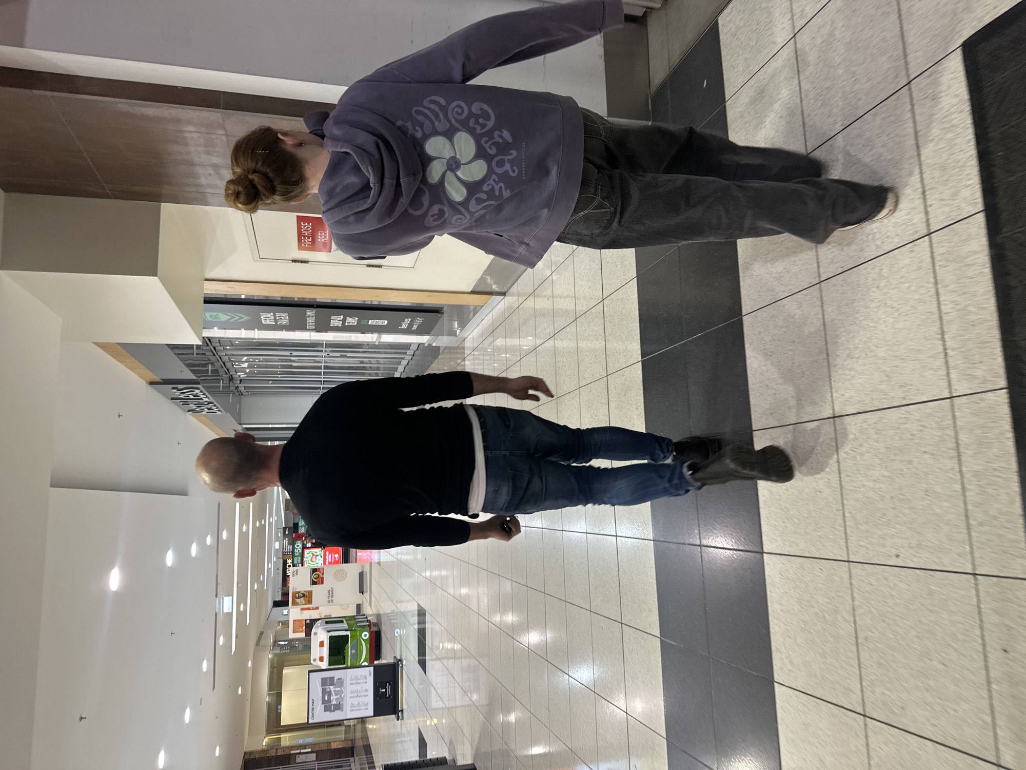 The image depicts the interior of a shopping center, likely located in Toormina, Australia, with a focus on the flooring and the backs of two individuals walking away from the camera. The floor is tiled with large square tiles, featuring a prominent alternating pattern of light beige and dark grey. The lighting appears to be artificial, typical of an indoor commercial space, casting reflections on the polished floor.
In the foreground, the lower legs and feet of two people are visible. The person on the left is wearing dark denim jeans and black boots, with the lower portion of their left leg prominently featured. The person on the right is wearing dark grey jeans and appears to be wearing sneakers.
The mid-ground reveals more of the individuals. The person on the left has a shaved head and is wearing a black long-sleeved shirt and blue jeans. The person on the right is wearing a purple hooded sweatshirt with a floral graphic design on the back, their hair is pulled up in a bun.
The background shows the architecture of the shopping center, including shopfronts with displays and signage. One sign reads "FIRE HOSE REEL." Further down the corridor, there are various retail displays and a small train-like ride. The environment suggests a public indoor space, possibly a mall or shopping precinct. The perspective is from behind the individuals, looking down the length of the corridor.