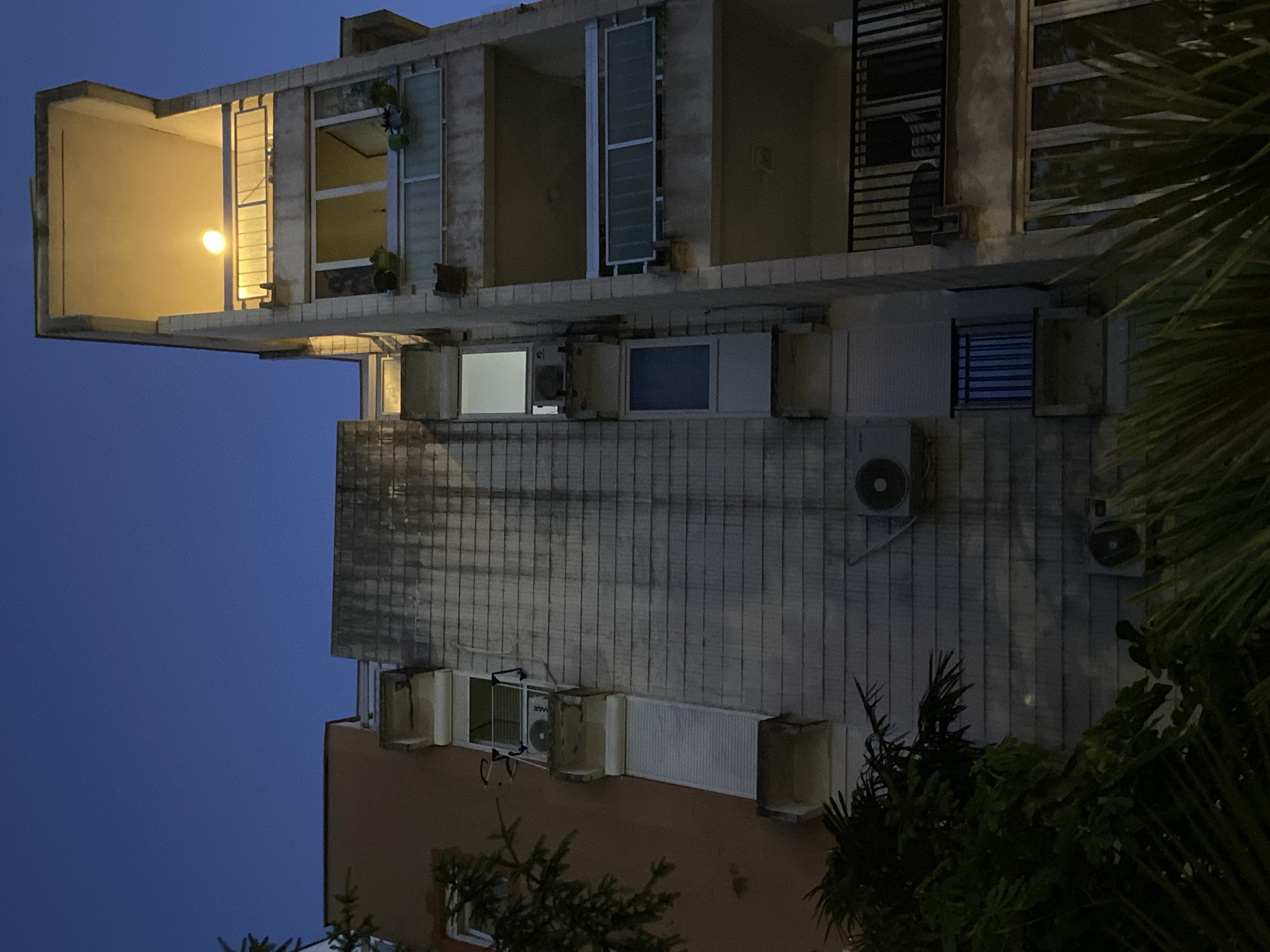 The image depicts an exterior view of a multi-story building at night in an unidentified location. The building features multiple windows and balconies with varying designs. Some windows have air conditioning units installed. The facade of the building is aged, with visible signs of wear and water streaks. A light is visible within one of the balconies. Dark foliage of nearby trees partially obscures the lower portion of the building and the right side of the frame. The sky in the background is a deep blue.