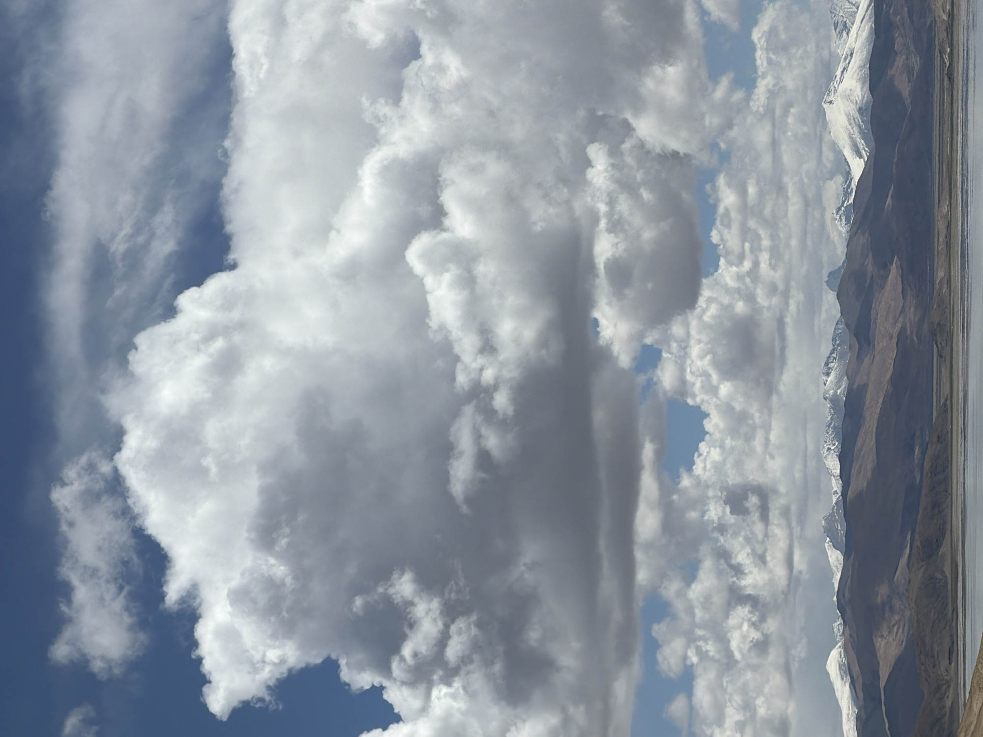 This landscape image captures a majestic, untamed natural scene, likely in the mountainous regions near Labrang township, China. The primary subjects are the vast sky, towering mountains, and a serene body of water.
The upper two-thirds of the horizontal vista are dominated by a dramatic sky filled with an array of clouds. Bright white, fluffy cumulus clouds are interspersed with larger, darker grey clouds, creating a dynamic atmospheric display. Patches of deep blue sky are visible, contrasting with the dense cloud cover.
Below the sky, a long, rugged mountain range stretches across the mid-ground. These mountains are predominantly earthy brown and grey in color, characterized by their steep slopes and sharp, imposing peaks. Many of the higher summits are capped with pristine white snow, which starkly contrasts with the darker rock, accentuating the rugged contours of the terrain. The snow indicates a high-altitude environment.
In the foreground, a calm body of water, possibly a lake or a wide river, extends along the base of the mountains, appearing still and reflective.
The scene is devoid of any human presence or man-made structures, emphasizing its natural grandeur and remoteness. The weather appears to be partly cloudy during the daytime, with ample light illuminating the landscape. The overall impression is one of vastness, tranquility, and the awe-inspiring beauty of high-altitude wilderness. No visible text is present in the image.