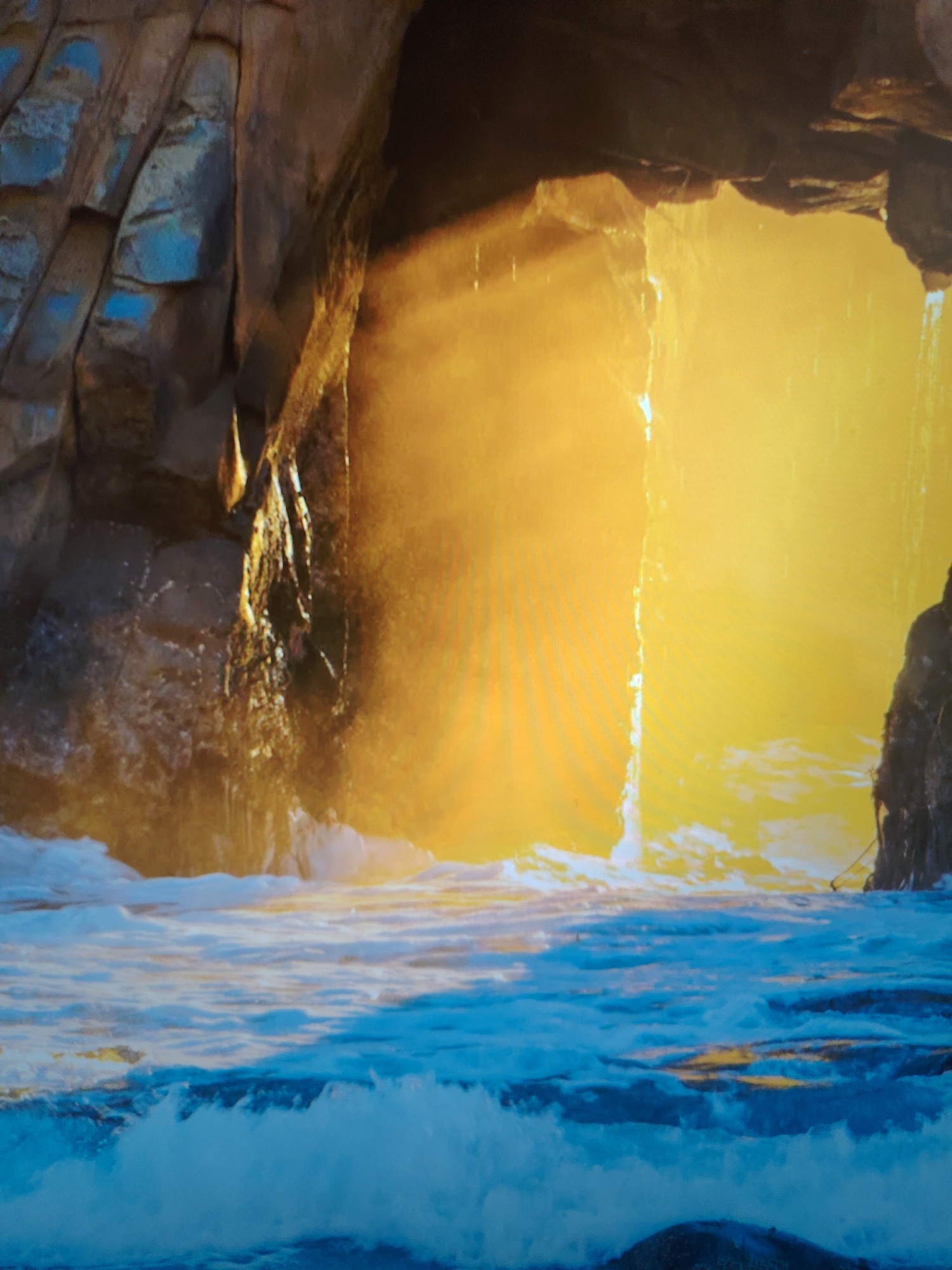 The image depicts a coastal scene with a large rock formation featuring a cave opening. The interior of the cave is brightly lit, possibly by sunlight. Water, indicated by waves and foam, is visible both inside and outside the cave entrance. The rock formation exhibits a layered structure and various textures, while the water has dynamic movement evident in the wave patterns.