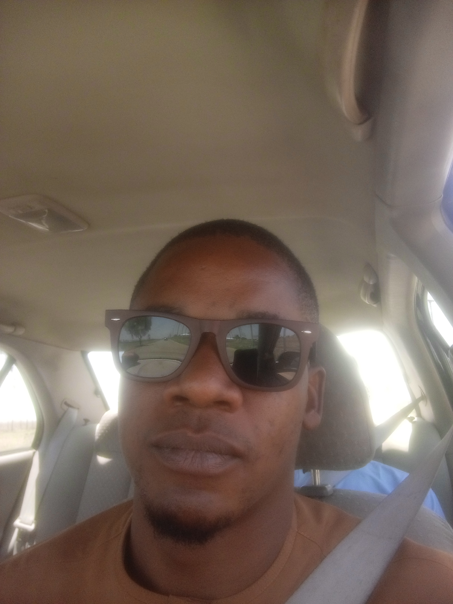 A man is in the front seat of a car. He is wearing sunglasses and a seatbelt. The car's interior is beige. The reflection in his sunglasses shows a road. The location cannot be determined based solely on the image.