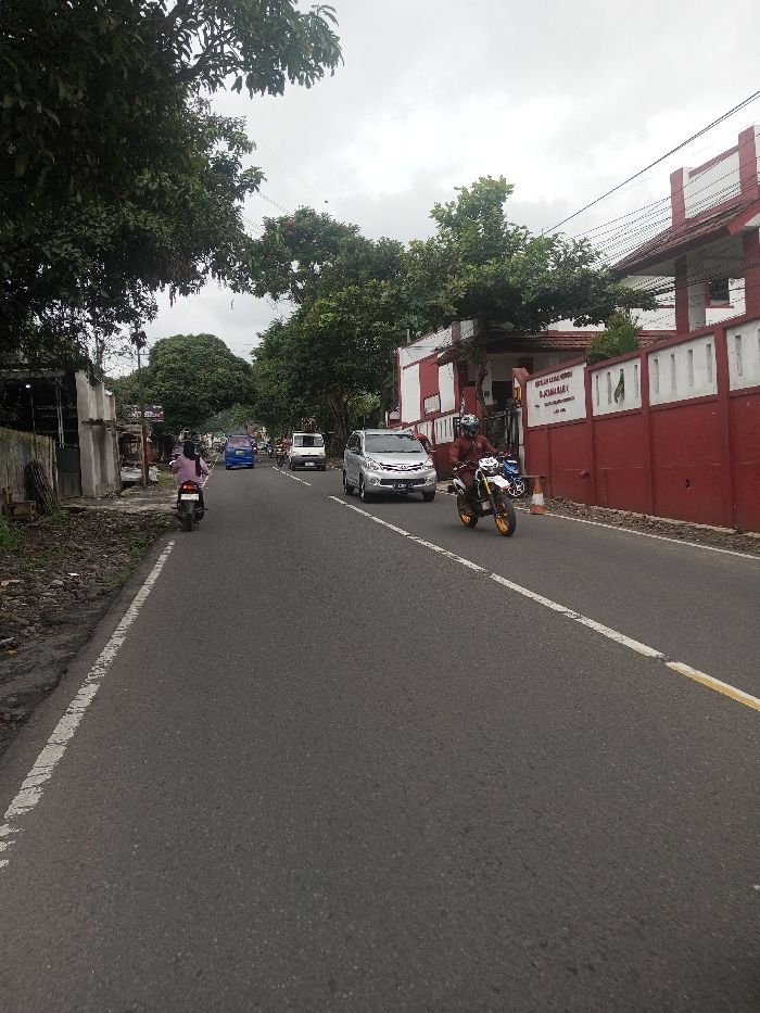 A street scene features a motorcycle approaching the camera, a silver car, and other vehicles further down the road. A woman on a scooter is on the left side of the road, heading in the opposite direction. Lush green trees line both sides of the street. A building with a red and white facade stands on the right, with a sign visible on its wall. The road is asphalt, with a yellow line on the right edge. The sky is overcast.