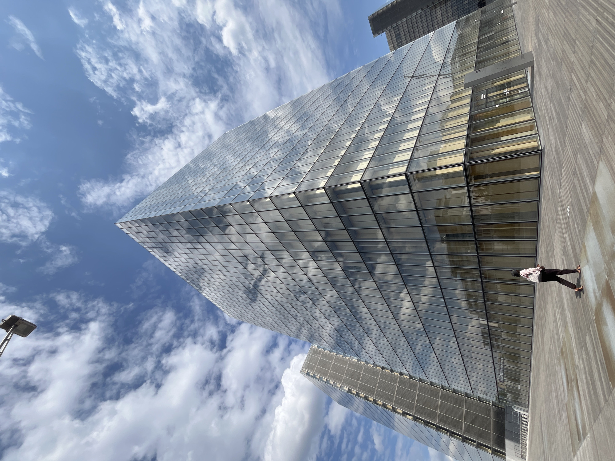 A person is walking on a concrete sidewalk next to a modern glass building with a reflection of clouds in the windows. The building is tall and angular, with a facade of glass panes. The sky is blue with scattered white clouds. A street light is visible on the left side of the image. The scene appears to be outdoors in daylight, likely in an urban environment. The person is wearing dark pants and a light-colored shirt and is walking away from the camera. The image is taken from a low angle, looking up at the building and sky. The lighting suggests it is daytime, and the weather is clear with some clouds.