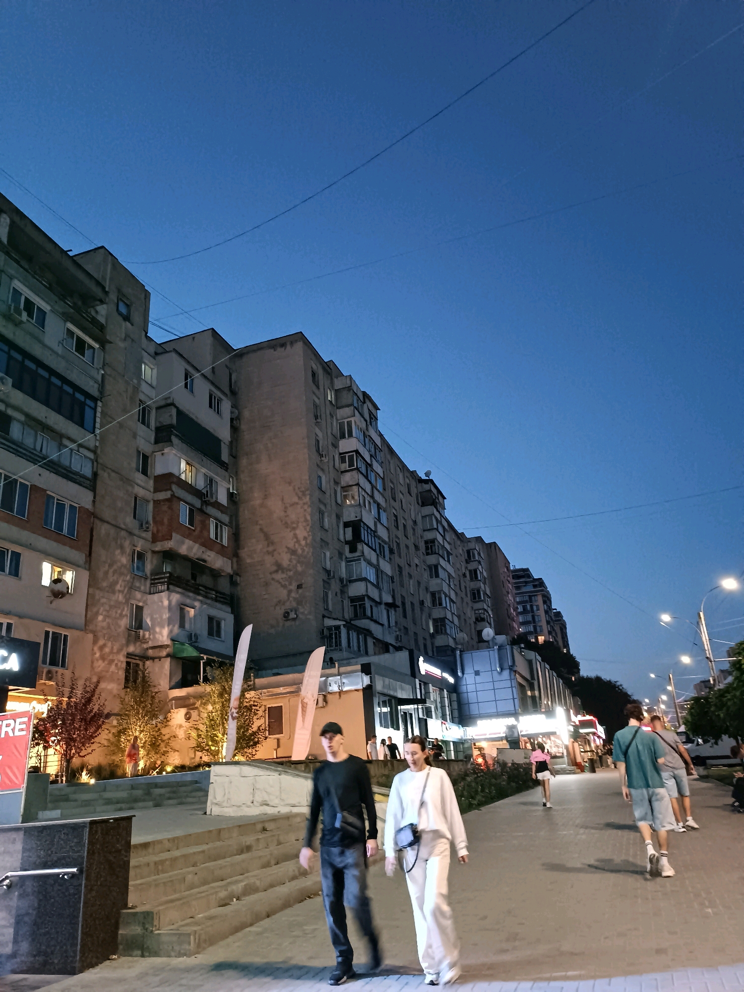 This is a full shot, vertical, outdoor, eye-level, medium-light photograph taken at dusk of a street scene in Chișinău, Moldova. In the foreground, a man and a woman are walking away from the camera on a paved sidewalk. The man wears a black long-sleeved shirt, dark pants, and a black crossbody bag. The woman wears a white hooded sweatshirt, white pants, and a black crossbody bag. Behind them, several other people are walking in the same direction, including two men in shorts and t-shirts and a person in a pink top and shorts. To the left of the sidewalk are stairs leading up to a building, with trees and lighting. The background features several multi-story apartment buildings with balconies and windows. Streetlights are visible in the distance. The sky is a deep blue, indicating twilight.