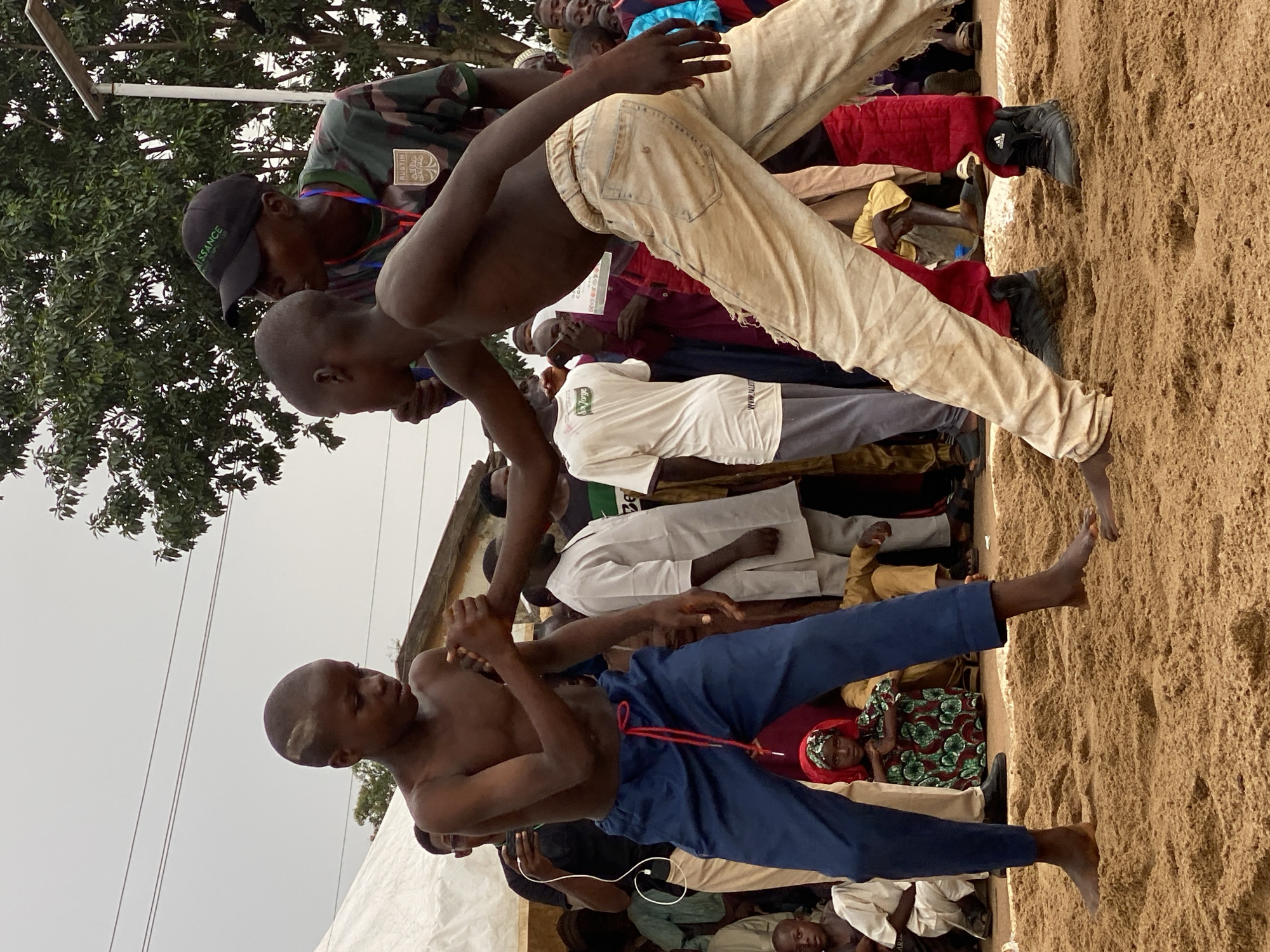 Two shirtless young men are wrestling in a sand pit in Kafanchan A, Nigeria. The wrestler closer to the viewer wears blue pants secured with a red drawstring and stands with his right foot firmly planted in the sand. His opponent is dressed in cream-colored pants and appears to be off-balance, poised on one foot. A referee wearing a camouflage shirt, black baseball cap, and lanyard stands above the wrestlers. Spectators surround the wrestling pit, including people dressed in traditional West African clothing. The event is held outdoors under an overcast sky. A few trees are visible.