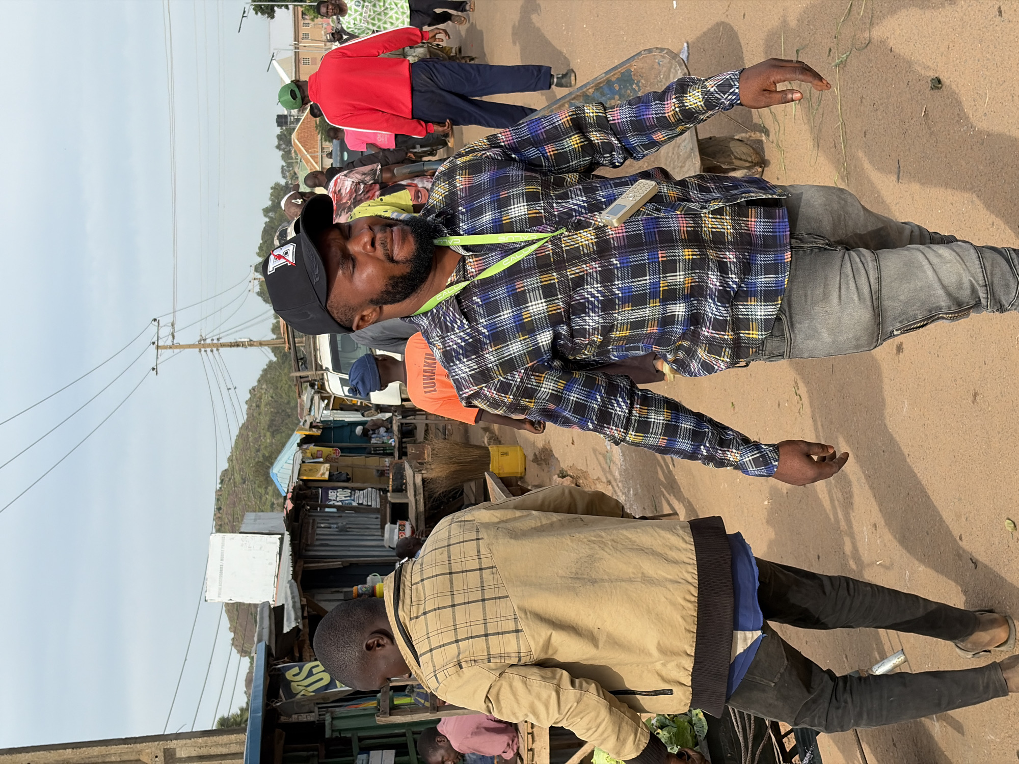 The image depicts a bustling street scene in Gura Top, Nigeria. In the foreground, two men are walking towards the viewer. The man on the right, closer to the camera, wears a black baseball cap with a white logo, a plaid shirt with blue, white, black, and yellow patterns, and grey jeans. A lanyard with a phone attached hangs from his neck. He has a beard and is looking downwards. The man on the left wears a tan plaid jacket over a blue garment and dark pants. He is looking to his left and appears to be carrying something that is not fully visible.
The background is filled with market stalls and people. Various shops with corrugated metal roofs and open fronts display goods. There are other individuals in the background, some walking, some standing, and some possibly engaging in commerce. The environment appears to be a dusty, outdoor market area with a cloudy sky overhead. Overhead wires and poles are visible, suggesting an urban or semi-urban setting. The overall atmosphere is one of active street life and commerce.