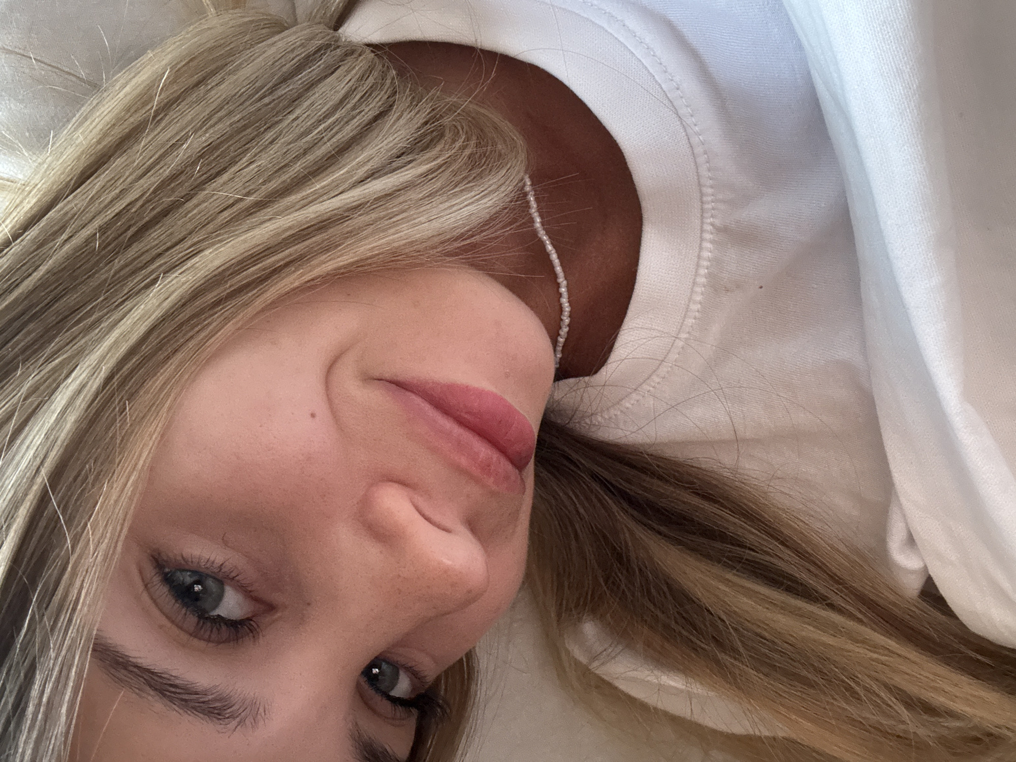This is a close-up, tilted selfie of a young woman with blonde hair. She is wearing a white t-shirt and a delicate pearl necklace. Her eyes are open and looking towards the camera, and she has a slight smile. The background is a soft, neutral color. There is no indication of the city of Sokhumi, Abkhazia in the image.