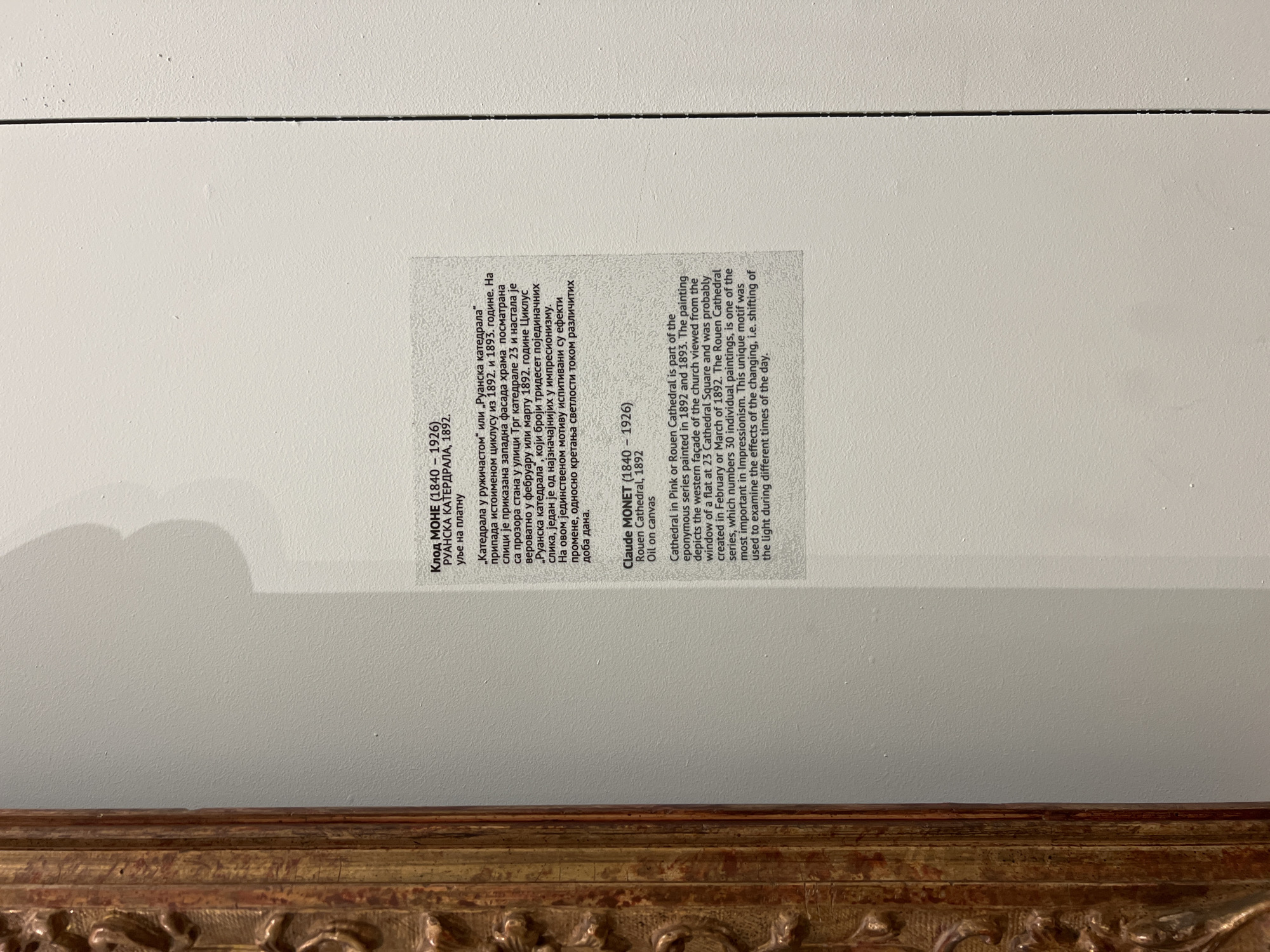 A close-up shot shows a wall-mounted descriptive plaque for a Claude Monet painting, likely displayed in a museum or gallery setting in Belgrade, Serbia. The plaque includes information in both Serbian and English. The Serbian section provides the artist's name (Клод МОНЕ, Claude Monet), the painting title (РУАНСКА КАТЕРДРАЛА, Rouen Cathedral), the year (1892), and the medium (уље на платну, oil on canvas). Below this, a paragraph describes the painting as part of the Rouen Cathedral series, detailing its depiction of the western façade from a window at 23 Cathedral Square and its exploration of the changing light. The English section repeats similar information. The plaque is mounted on a white wall, visible above a portion of an ornate, gold-toned picture frame.