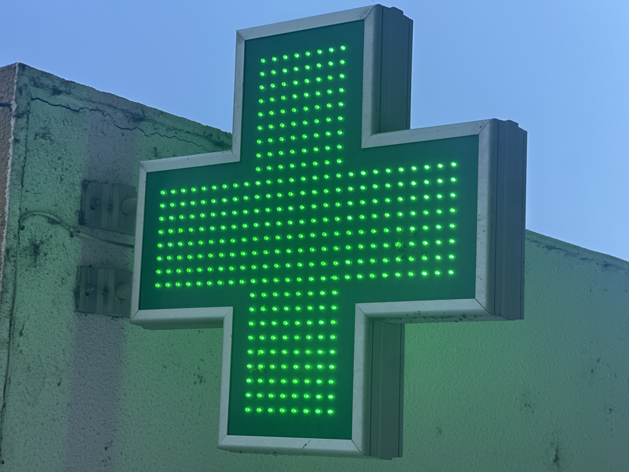 The image shows a close-up of a bright green, LED-lit pharmacy cross sign against a clear blue sky. The cross is the primary subject, made of numerous small, evenly spaced green lights forming its shape. The sign appears to be mounted on the exterior of a building, partially visible on the left and right sides of the frame.
To the left of the sign, a section of the building's exterior wall is visible. This wall has a rough, textured surface and shows signs of wear, including a prominent crack running along the upper edge. There are also metallic mounting brackets attached to this wall, suggesting the sign is attached to it.
To the right of the sign, the building continues, with its textured surface also visible. The sign itself has a light-colored frame, likely metal or plastic, which surrounds the green LED display.
The lighting suggests it is daytime, with the clear blue sky indicating fair weather. The bright illumination of the green LEDs suggests the sign is functioning and visible, possibly as a nighttime indicator or a permanent fixture. The overall impression is of a functional, albeit slightly aged, urban or commercial environment. There is no visible text in the image.