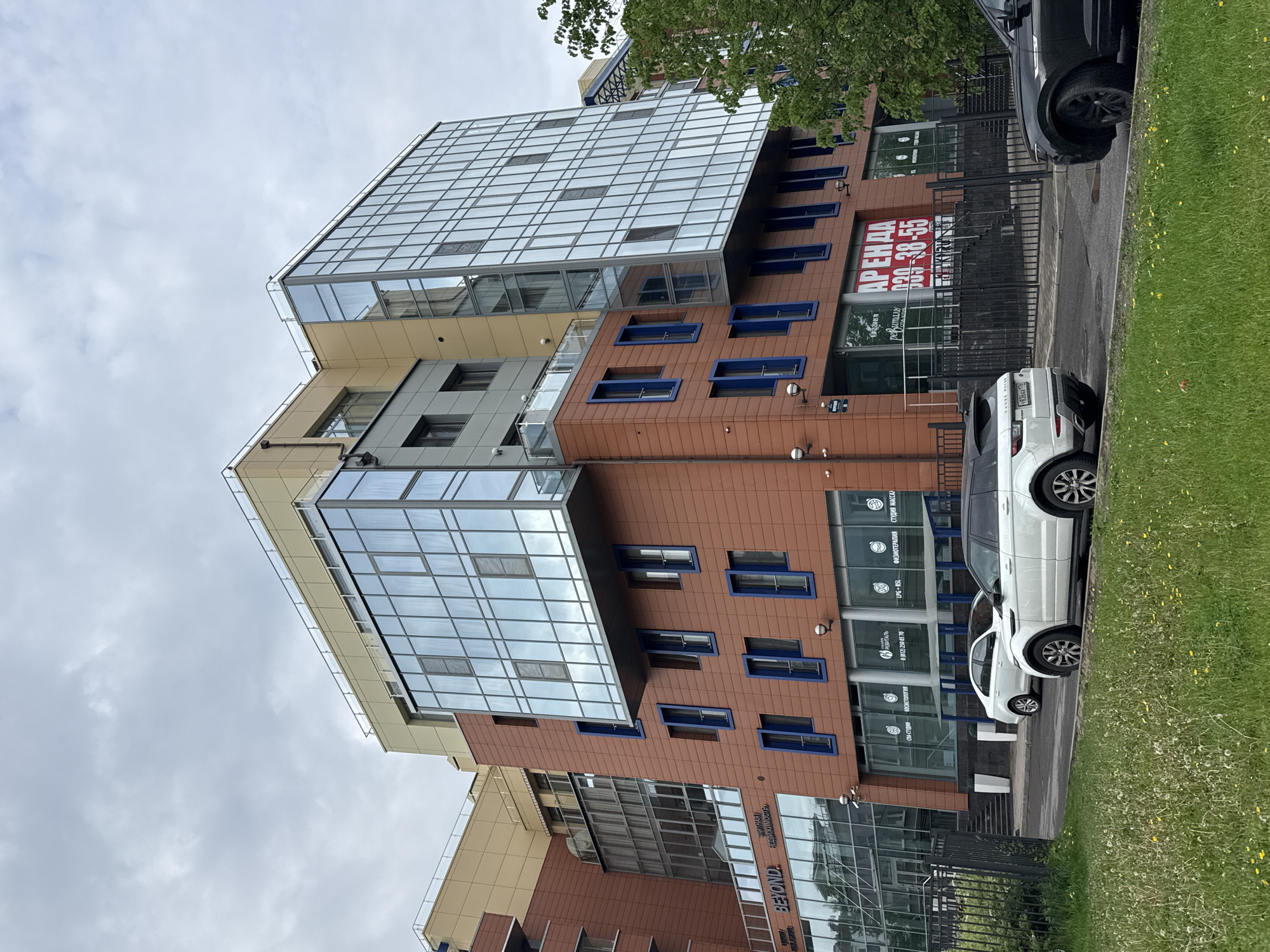 A multi-story building with a modern architectural design stands adjacent to a grassy area and a parked white vehicle. The building features a combination of brown tile facade and reflective glass windows with blue frames. A sign in Russian script, prominently displayed on the building, advertises rental spaces with a phone number. Part of a dark-colored car is visible at the top of the frame near the building. The sky is overcast. The building is located in the city of Saint Petersburg, Russia.